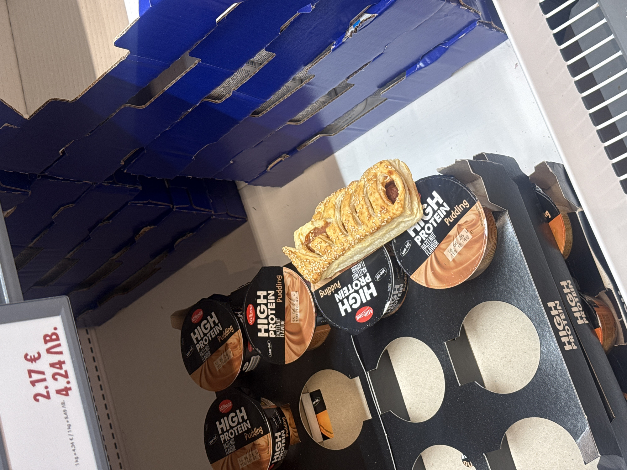 The image shows a shelf in a store, possibly a supermarket in Sofia, Bulgaria, where a package of "Milbona High Protein Hazelnut Flavor Pudding" is stocked. The price tag on the left shows "2.17 €" and "4.24 ΛΒ." (likely Bulgarian Lev), indicating the pudding's cost in both Euros and the local currency. A pastry is placed atop the pudding cups. The pudding is in a cardboard container with circular cutouts to hold individual cups. The packaging is predominantly black with white and brown accents. The shelf above is constructed with corrugated cardboard painted blue.
