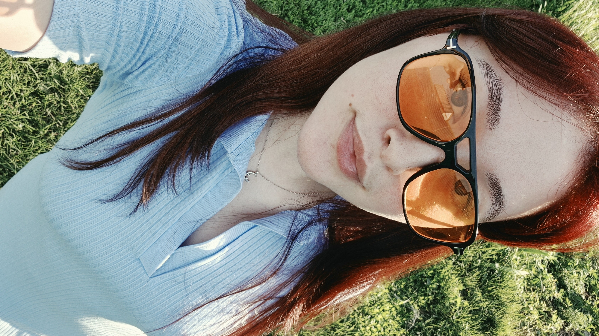 A close-up, overhead shot shows a woman with auburn hair lying on green grass. She is wearing large black sunglasses with orange lenses, and a light blue collared shirt. A silver necklace with a crescent moon pendant is visible around her neck. The woman's face is slightly tilted, and she is looking towards the camera. The sun is casting shadows on the grass and the woman's face.