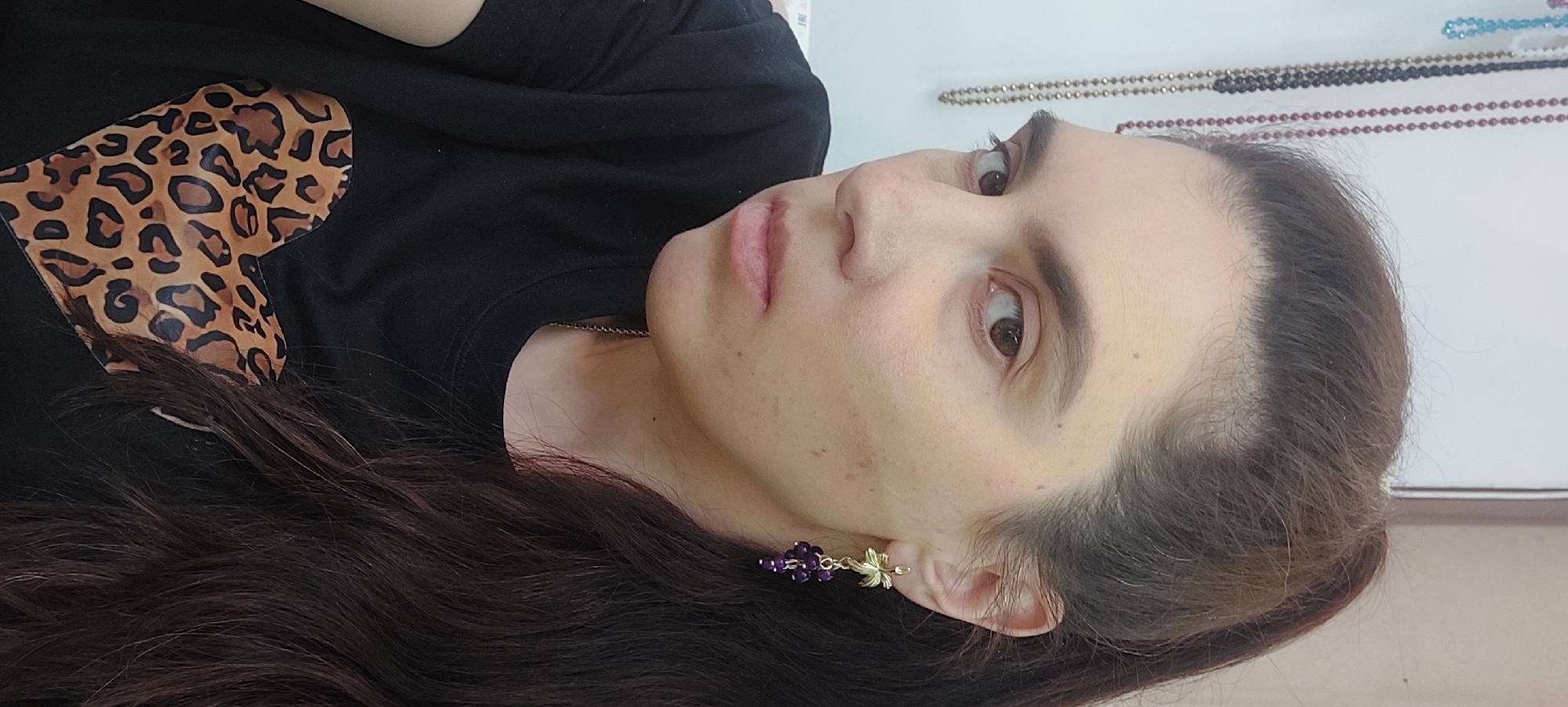 A woman with dark hair is shown in a close-up shot, angled slightly to the side. She is wearing a black T-shirt with a leopard print heart on it. She has light skin, brown eyes, and a neutral expression. She is wearing an earring that appears to be purple and gold. The background is out of focus but appears to be a room, possibly with a white wall. There are strands of beads hanging in the background. There is no information to definitively place this image in Novosibirsk, Russia.