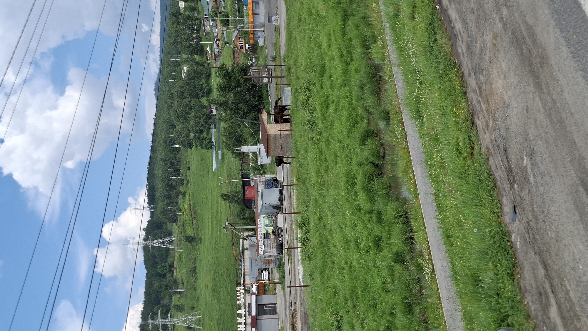 The image shows a rural or semi-rural area, possibly on the outskirts of a town. In the background, there is a green, rolling hill covered in trees and dotted with some power lines and transmission towers. A few buildings and houses are visible scattered on the hillside and at the bottom of the hill. In the mid-ground, there are some industrial or commercial buildings with signage. A paved road is visible in the foreground, with a strip of green grass and a small stream or ditch separating it from the road. There are several horses standing near a small wooden structure, possibly a stable or shelter. The sky is blue with scattered white clouds, and power lines are strung across the top of the image. The scene appears to be set in a natural environment with evidence of human infrastructure.