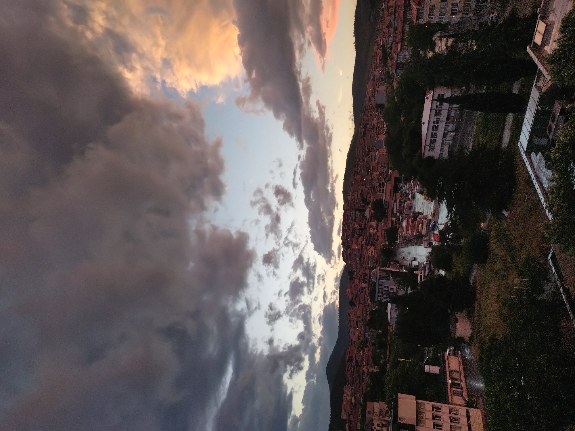 The image captures a twilight cityscape in Stara Zagora, Bulgaria, with the view seemingly taken from an elevated position, possibly a balcony or rooftop. The foreground is dominated by dramatic, multi-layered clouds that are transitioning from dark gray to lighter tones, with touches of orange and pink highlighting the upper edges.
In the middle ground, a hillside is covered densely with red-tiled roofs of residential buildings, typical of urban landscapes. Scattered among the residential structures are larger white-colored buildings that are interspersed with greenery.
The foreground to the right shows buildings with architecture ranging from low-rise to high-rise, obscured by trees.
The time appears to be dusk, as evidenced by the soft light on the buildings and the cloud formations in the sky. The overall impression is one of urban density juxtaposed with natural elements.