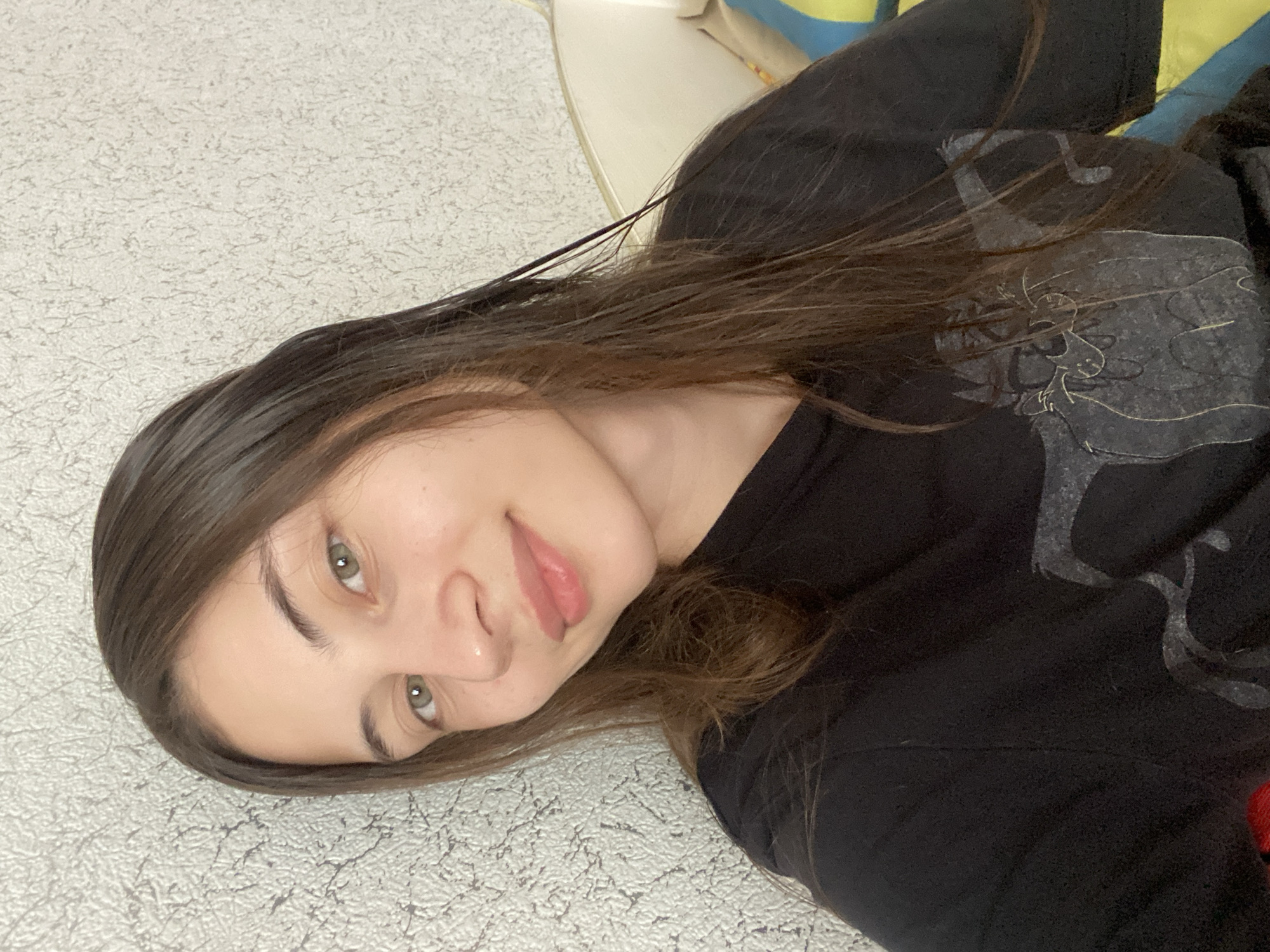 A young woman with long, dark brown hair and green eyes is looking directly at the camera. She is wearing a black t-shirt with a graphic print of a cat. The background consists of a textured, light-colored wall and a blurred section of a yellow and blue striped object. The image appears to be a selfie taken indoors. The specific location within the city of Votkinsk, Russia, cannot be determined from the visual information provided.