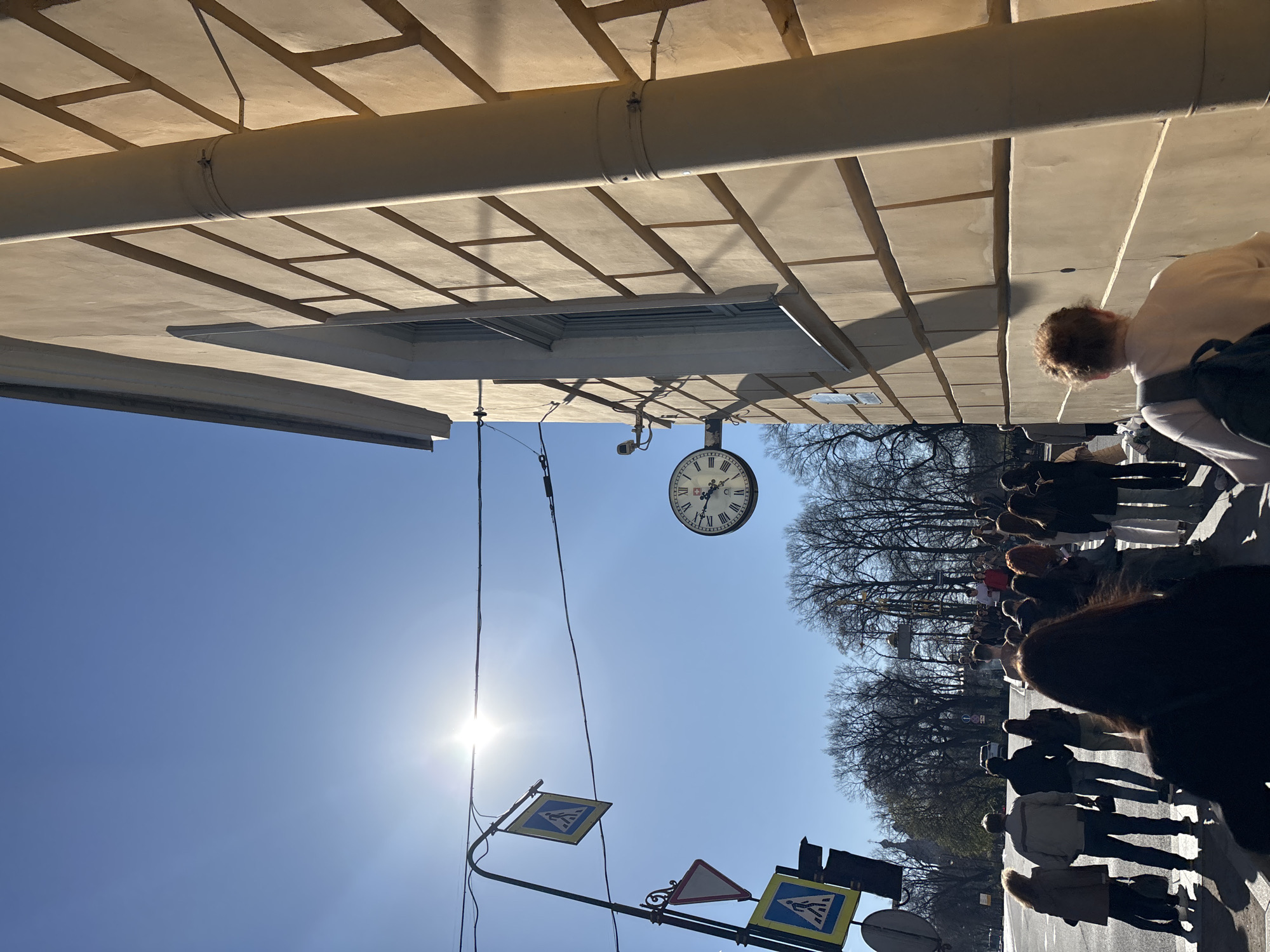 The image shows a street scene in Saint Petersburg, Russia. The photograph is taken at an oblique angle, pointing upwards. The sky is bright blue with the sun shining. A building overhangs with a pipe running along the bottom. A clock is attached to the building. Beneath the clock are a set of traffic and pedestrian crossing signs. People are walking on the sidewalk, lining up to cross the road. Bare trees line the background.