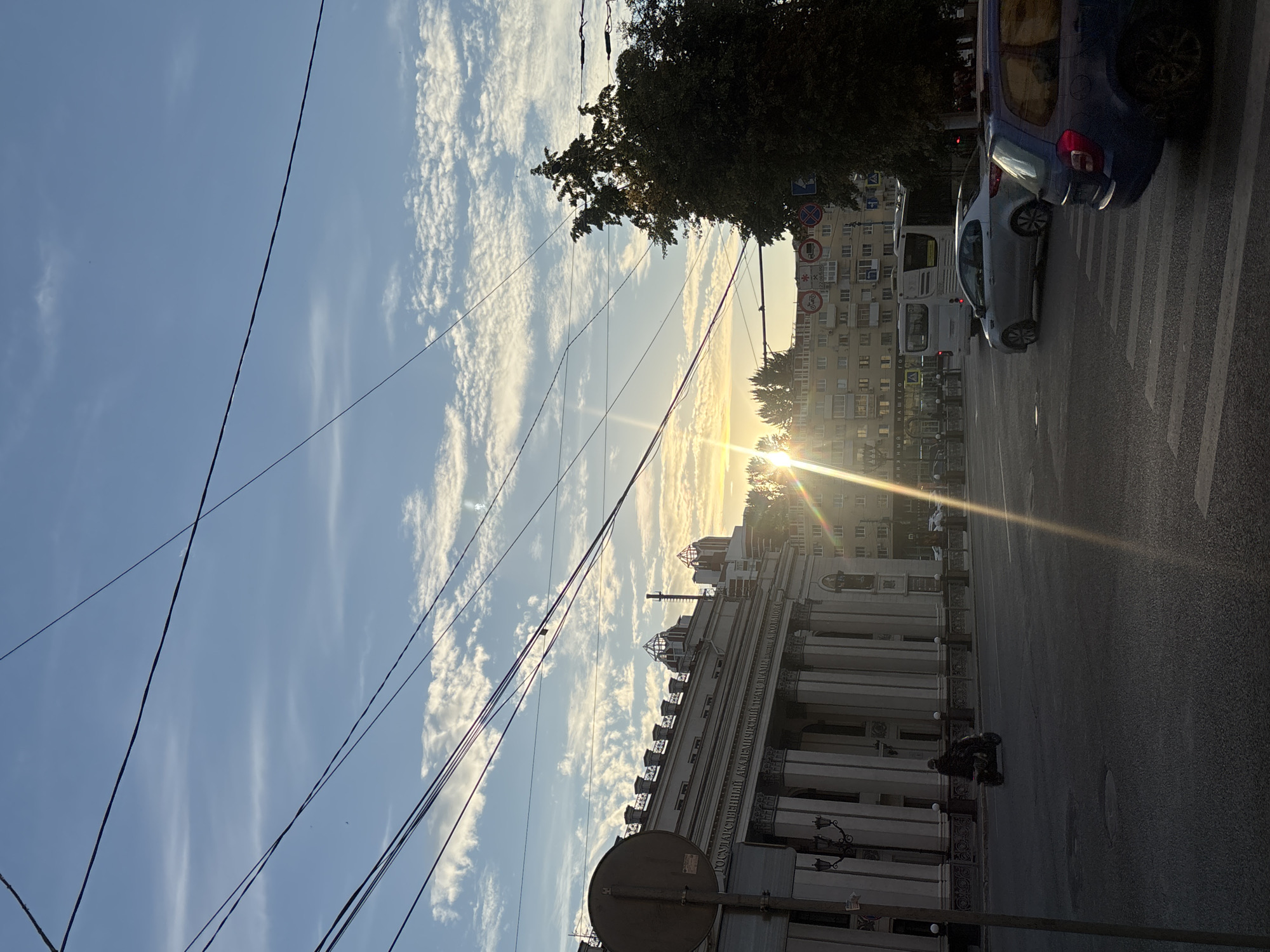 This media file depicts an urban scene in Novaya Usman', Russia, during what appears to be late afternoon or early evening, indicated by the low angle of the sun and the warm, golden light casting long shadows. The sky is a clear blue, dotted with fluffy white clouds, suggesting fair weather.
The image captures a street with a pedestrian crossing marked by white stripes on the asphalt. Several vehicles are present, including a blue van, a silver car, and a white bus, suggesting active traffic. In the foreground, a building with classical architectural elements, featuring columns and ornate detailing, is visible. Text on the building reads: "ГОСУДАРСТВЕННЫЙ АКАДЕМИЧЕСКИЙ ТЕАТР ПУКИНИ" and below it "ИМ. А. КОЛЛОНТАЯ".
Overhead, a network of power lines crisscrosses the frame, adding a layer of urban infrastructure to the scene. A large tree with dense foliage is visible on the right side of the frame, partially obscuring the sun. A person on a scooter or motorcycle is seen on the right side of the road, near the building. Traffic signs are also visible near the tree. The overall mood is serene, with the setting sun illuminating the sky and the urban environment.