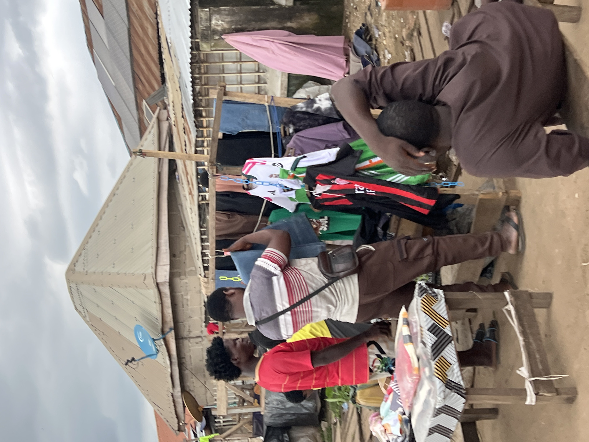 In Oko Amakom, Nigeria, an outdoor market scene unfolds under a cloudy sky. Three men are present. One man, wearing a red striped t-shirt, stands to the left, facing right. Another man, in a striped grey, red, and white t-shirt and brown cargo pants, stands in the center, holding up a pair of blue denim jeans. He has a dark shoulder bag. A third man, wearing a dark brown robe, is seated on a low wooden stool at the right, facing away from the camera, with his hand on his head.
In the foreground, a small wooden table covered with a patterned cloth displays folded items wrapped in clear plastic. Behind the people, a makeshift stall displays an assortment of clothing, including shirts and jerseys, hanging on a wooden frame. Various types of roofs, some made of corrugated metal and others with solar panels, are visible in the background, along with simple building structures. The ground is dirt.