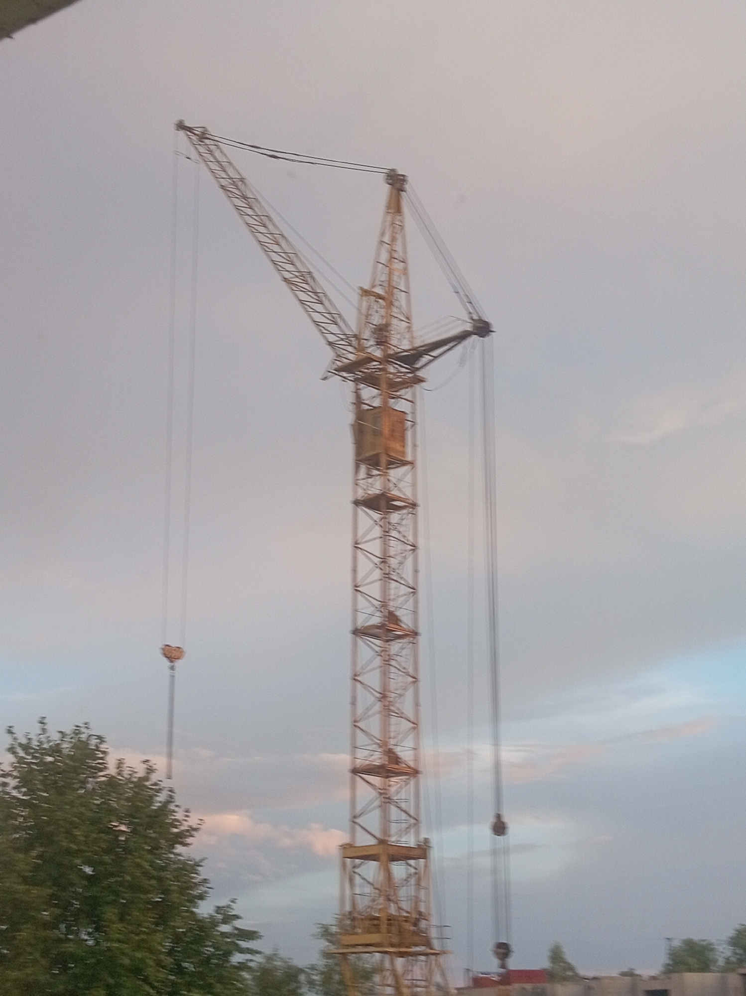 A tall yellow construction crane stands against a cloudy sky. The crane is a prominent structure, with its lattice boom extending upwards and to the left. A load block is visible suspended from the main cable. To the left of the crane, a lush green tree is partially visible, its branches extending upwards. In the distance, some buildings and more trees can be seen. The overall scene suggests an active construction site or an area undergoing development in the city of Shumilina, Belarus.