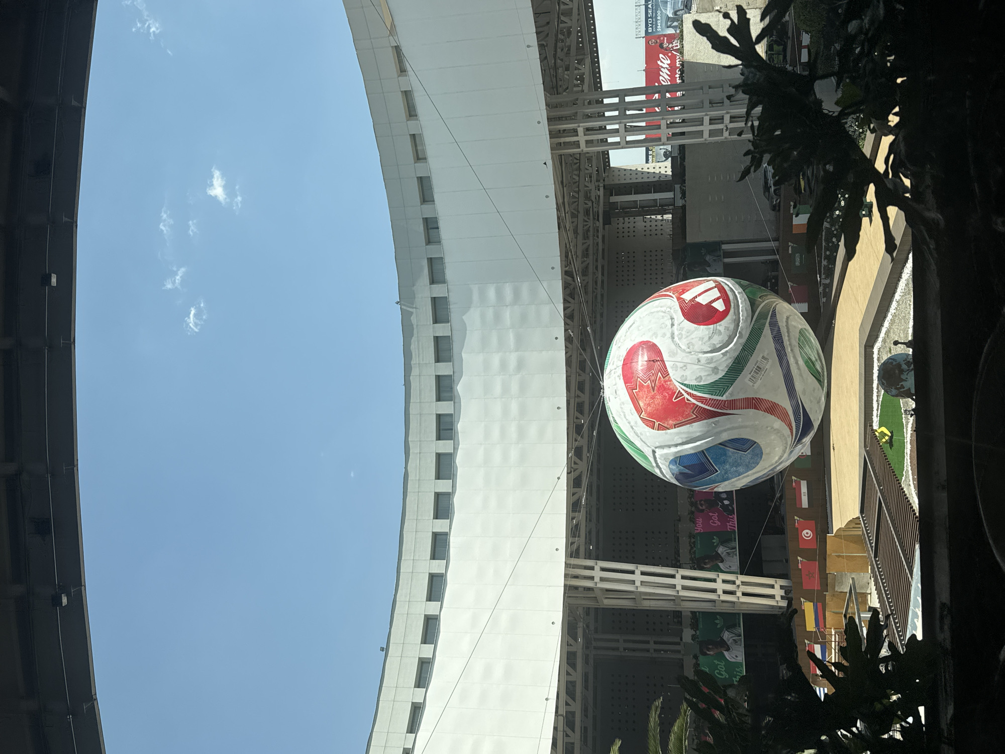 This image captures an outdoor scene, rotated clockwise, showcasing a large, inflatable soccer ball as the central subject, suspended in mid-air. The ball features a colorful design with red, white, blue, and green patterns, along with an Adidas logo.
On the left, a tall, multi-story building with numerous rectangular windows forms a prominent part of the background, its light-colored facade contrasting with the clear, bright blue sky above, which contains a few small, wispy clouds. Structural elements, including white metal beams and supports, are visible both near the building and around the hanging ball, suggesting a large public space or venue.
Below and to the right of the soccer ball, various national flags are displayed, including those of Tunisia, Morocco, and Colombia, indicating an international event. Banners featuring a person (likely a soccer player), an Adidas logo, and the motivational phrase "You Got This" are also visible. Dark foliage and plants frame the right side of the image. Further in the background, a large billboard partially displays "BYD SEAL" and "$949,800," along with the word "ente." A small patch of artificial turf with a miniature soccer goal and a small figure can be glimpsed below the ball.
The scene depicts a festive atmosphere, likely a public celebration or promotion related to soccer, possibly a World Cup or major tournament given the international flags. The bright daylight and clear sky suggest it is midday on a sunny day. The location is Mexico City, Mexico.