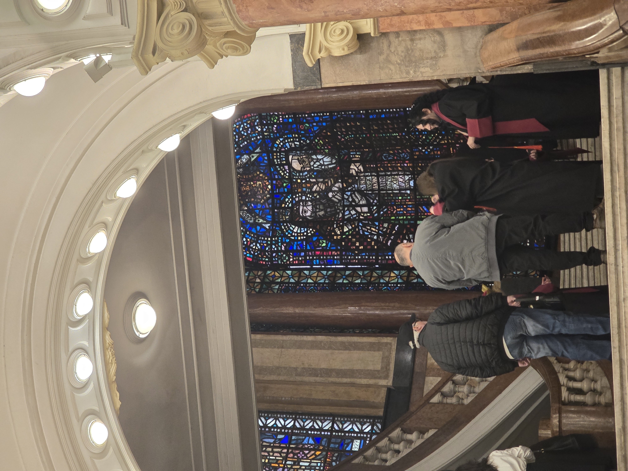 The image shows the interior of a church, likely in Sofia, Bulgaria based on architectural style. Several people are present, including two dressed in what appears to be religious robes. Other individuals are wearing casual clothing. A large stained-glass window is visible in the background. The architecture features arches, columns, and decorative molding. The light fixtures appear to be circular. Some people are standing on stairs, while others are near the stained-glass window. The overall impression is a visit inside an ornate church.