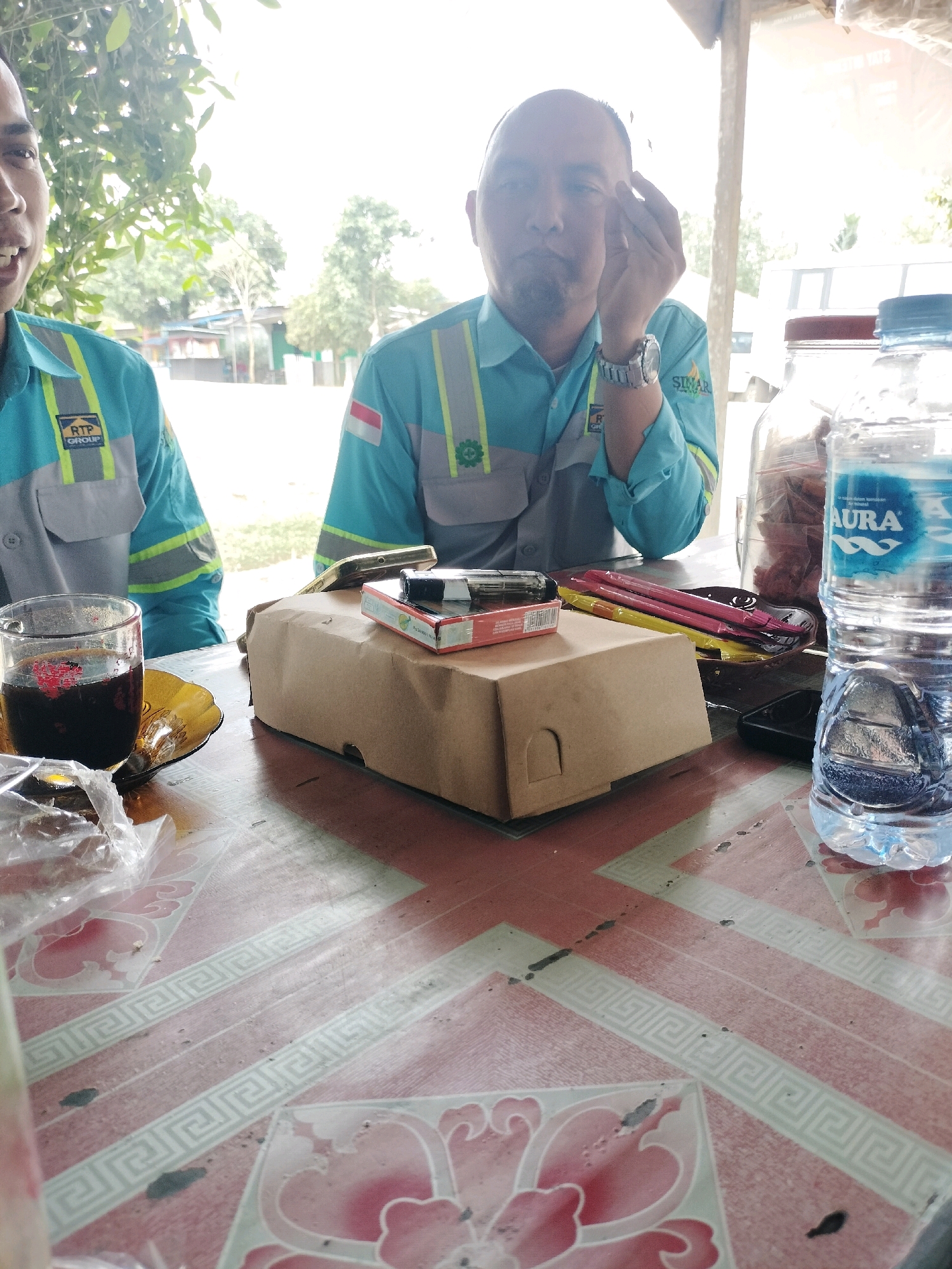 Two men in teal safety vests with reflective stripes are seated at a table outdoors, in what appears to be a break area. The man on the left is partially visible from the chest up, and the man on the right is visible from the shoulders up, with his hand near his face. In the foreground, a glass of dark liquid, a small plate with a piece of food wrapped in plastic, a cardboard box, a pack of cigarettes, and a bottle of Aura brand water are on the table. The table surface has a red and white patterned design. The background shows vegetation, buildings, and a bright sky. The scene is likely located in Maburai, Indonesia, given the presence of Indonesian flags on the uniforms.