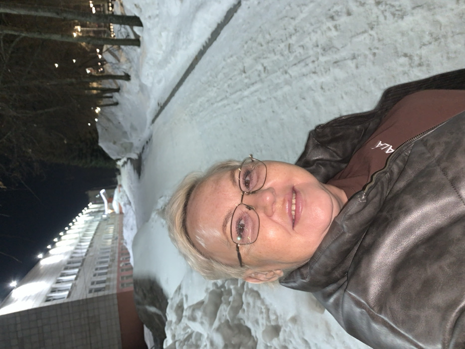 This image depicts a nighttime outdoor scene in Chaikovsky, Russia, under conditions of extensive snow cover. In the foreground, a woman with short, light-colored hair and gold-rimmed eyeglasses is captured from the chest up, smiling directly towards the camera. She is wearing a dark, possibly grey or dark brown, padded jacket over a brown shirt with partial white text, "ALÁ", visible on the right side.
The background on the left features a multi-story building with a light-colored facade, numerous rectangular windows, and a reddish-brown foundation. This building is brightly illuminated by a series of upward-facing floodlights positioned along its roofline. Significant snowdrifts are visible on the ground around the building. To the right, a snow-covered pedestrian path or cleared track extends into the distance, curving slightly. A line of bare deciduous trees stands along the right side of this path, with additional snow piled up alongside them. The sky is dark, confirming a nocturnal setting, with artificial light sources providing the primary illumination for the visible areas.