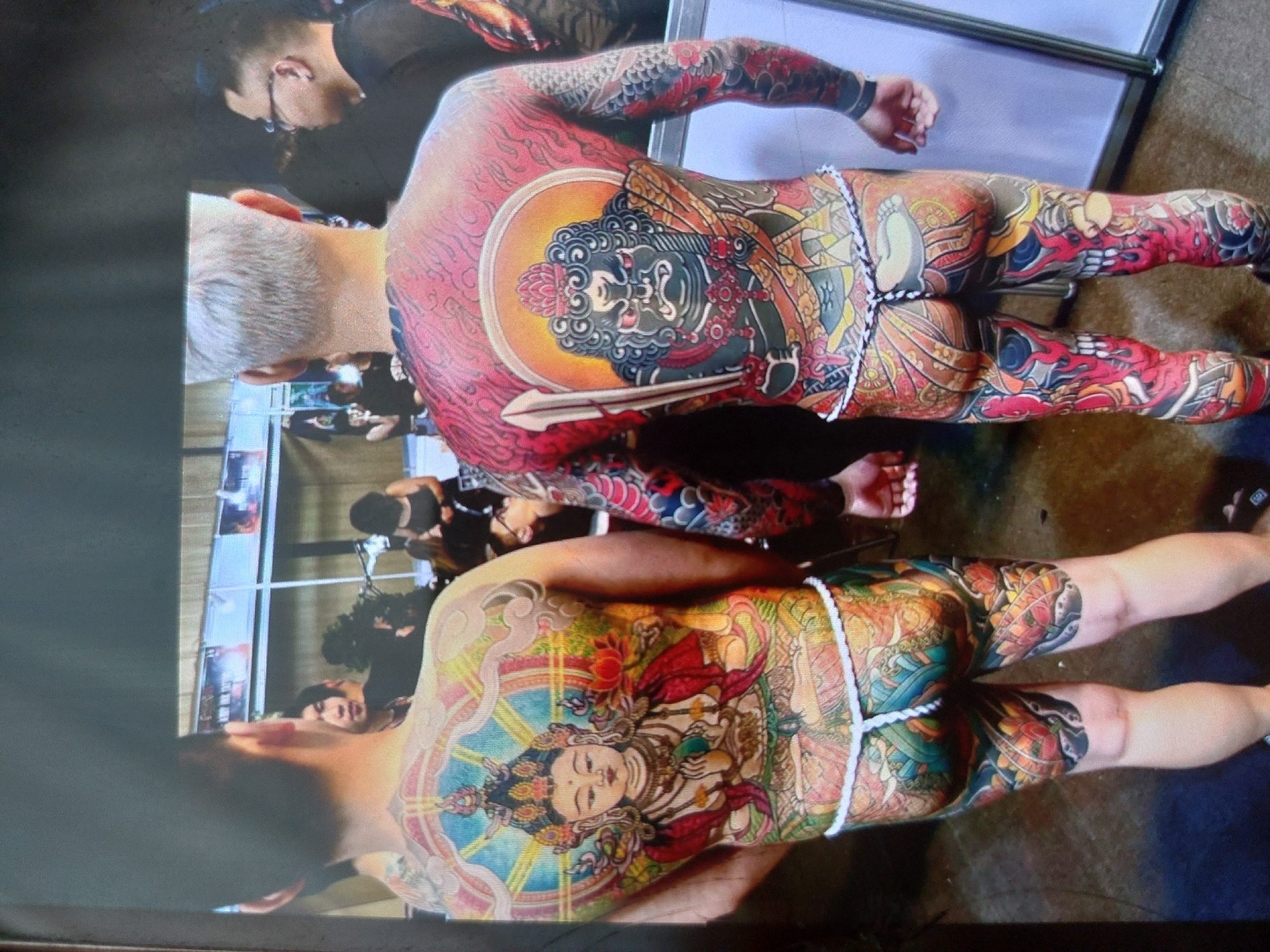 The image depicts two shirtless men standing close to each other. Both men have extensive traditional Japanese-style tattoos covering their backs, arms, buttocks, and legs. The man at the top has a red-toned tattoo with a dark-faced deity in a circular frame on his back, while the man at the bottom has a multicolored tattoo with a serene-faced deity. They are both wearing fundoshi (traditional Japanese undergarments), secured with white ropes.
The background includes a crowd of people, indoor settings, metal barriers, and posters. The location is not specified in the image. The city of Tsarevo, Bulgaria, is not relevant to this image.