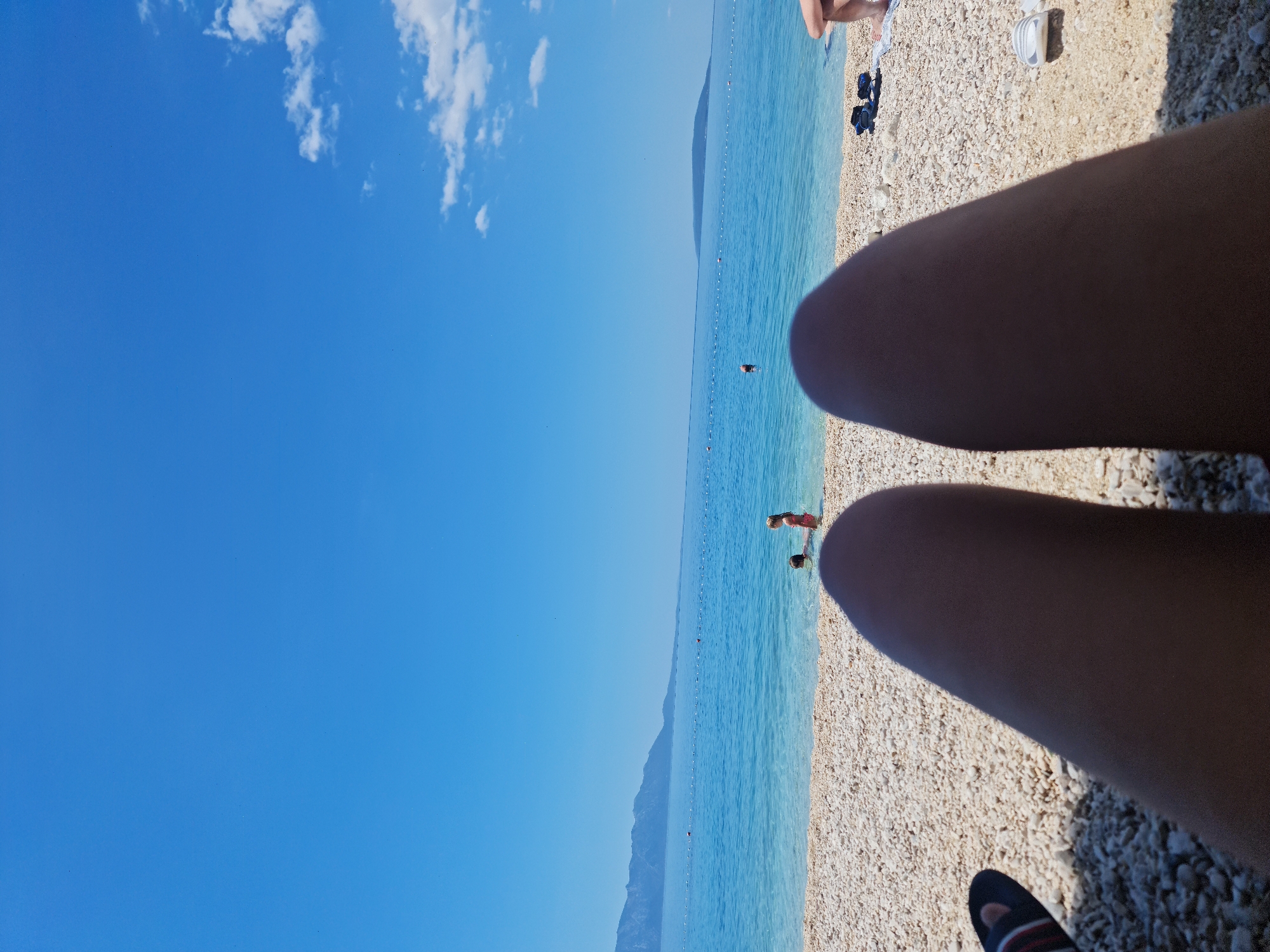 The image depicts a scenic view of a pebble beach on a sunny day. In the foreground, two legs of a person are visible, casting a shadow on the white pebbles. A pair of flip-flops with red and white stripes is placed to the left of the legs. The turquoise water of the Adriatic Sea stretches out towards the horizon, with a distant mountain range visible in the background. A few buoys mark a swimming area in the sea. In the shallow water, two children are playing, with one child appearing to be a girl in a pink swimsuit and another person partially submerged. The sky is a clear, vibrant blue with a few wispy white clouds on the right side. The location is identified as Općina Gradac, Croatia, a coastal municipality known for its beautiful beaches and clear waters.