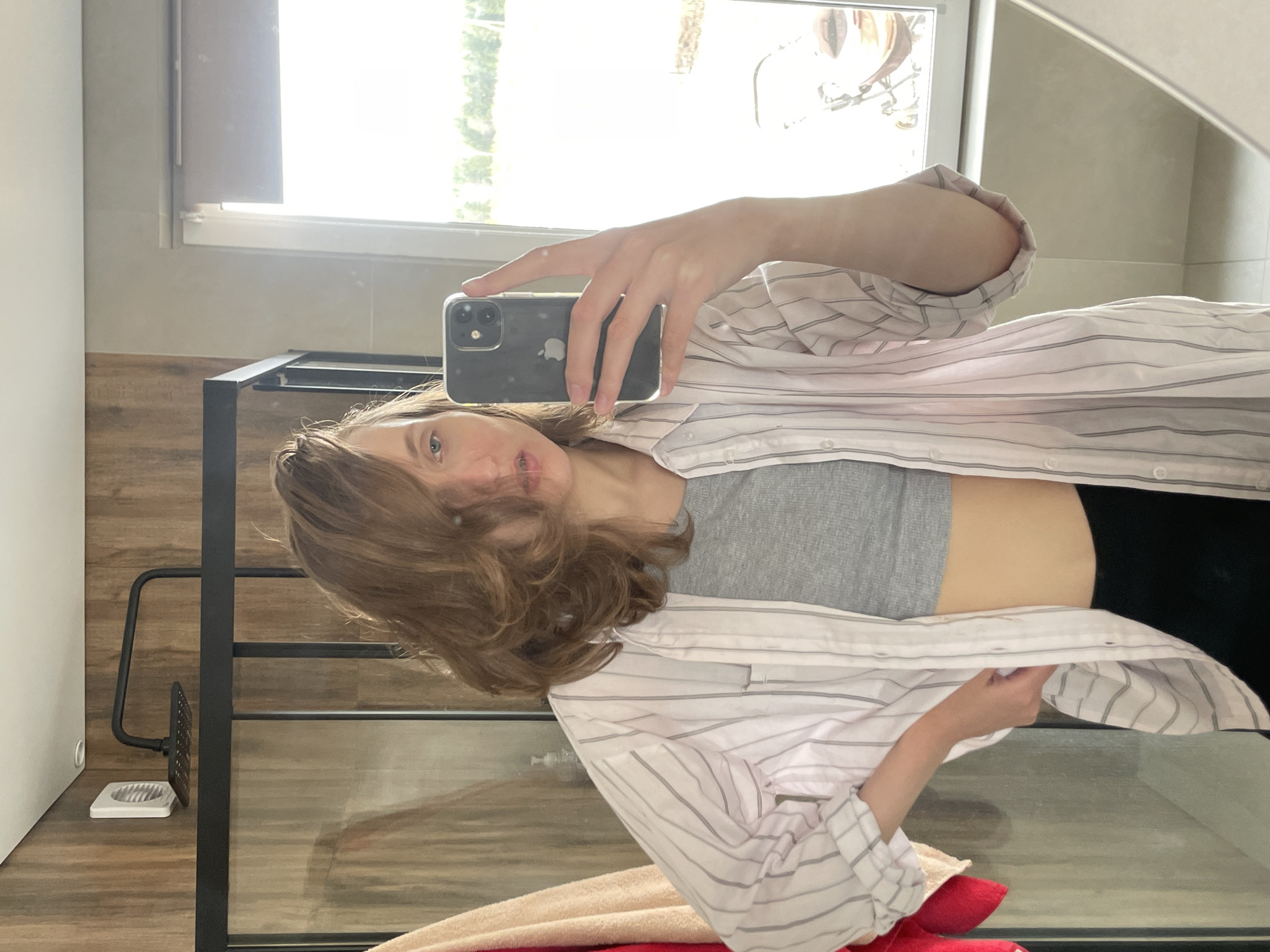 The image shows a young woman in a bathroom setting, possibly taking a selfie in a mirror. She is wearing a grey top and a white striped button-down shirt. She is holding a smartphone in her right hand. Behind her is a window with a view of trees. The location is unknown and cannot be deduced from the image.