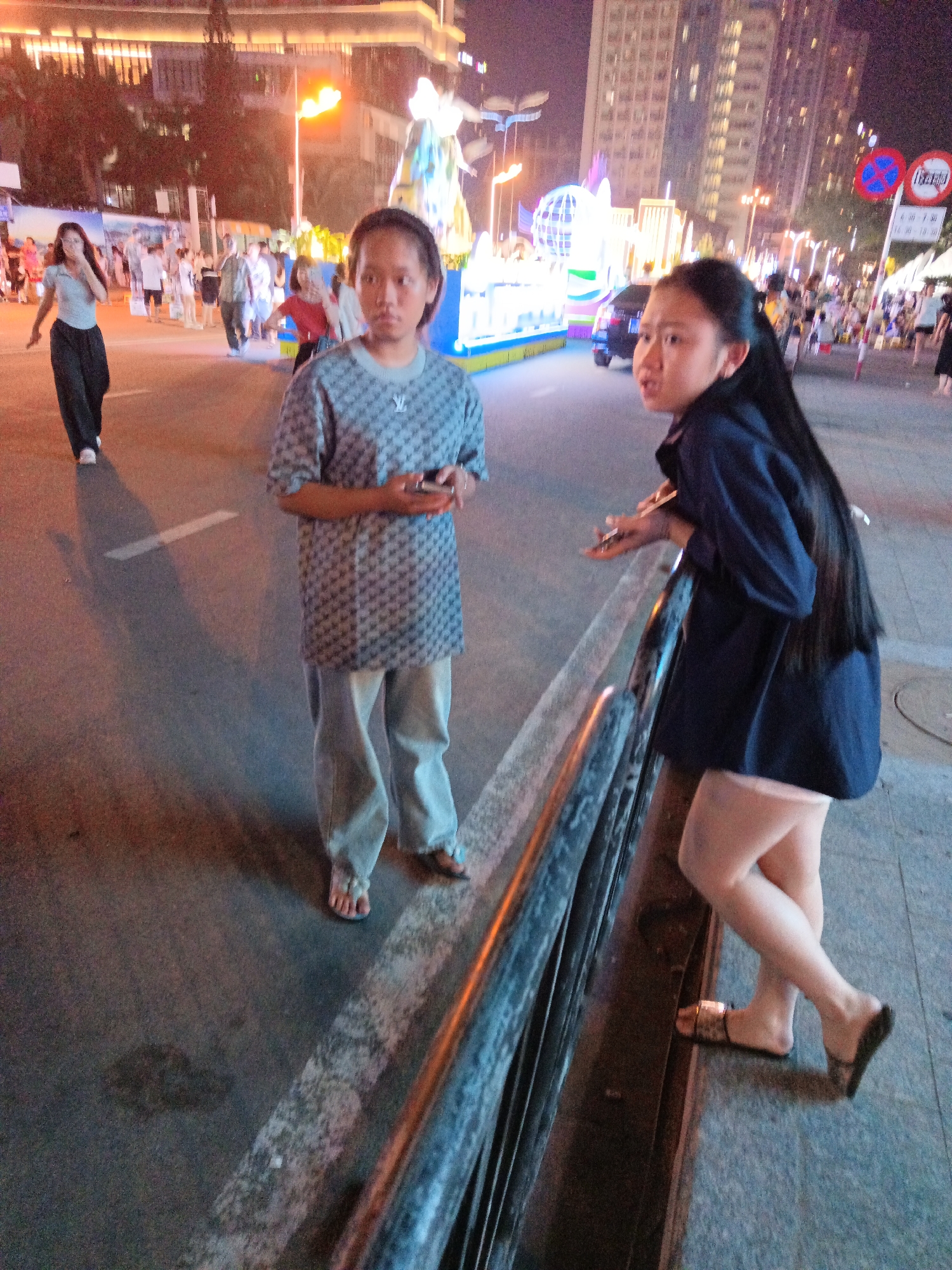 The image shows a street scene at night in Nha Trang, Vietnam, with a focus on two young women and the surrounding environment. The woman on the left is wearing a grey t-shirt with a repeating pattern of what appears to be LV monograms and light-colored pants. She is holding a smartphone in her hands. The woman on the right is wearing a dark blue jacket and light-colored shorts. She is leaning against a metal railing and appears to be looking to her left.
In the background, there are streetlights, tall buildings with illuminated windows, and decorative lights, suggesting a festive or commercial area. The street is paved, and there is a white line marking a lane. Several other people are visible in the background, walking and interacting. There is also a car and some decorative structures, possibly part of a public display or event. The overall atmosphere is lively and urban, illuminated by artificial light.
