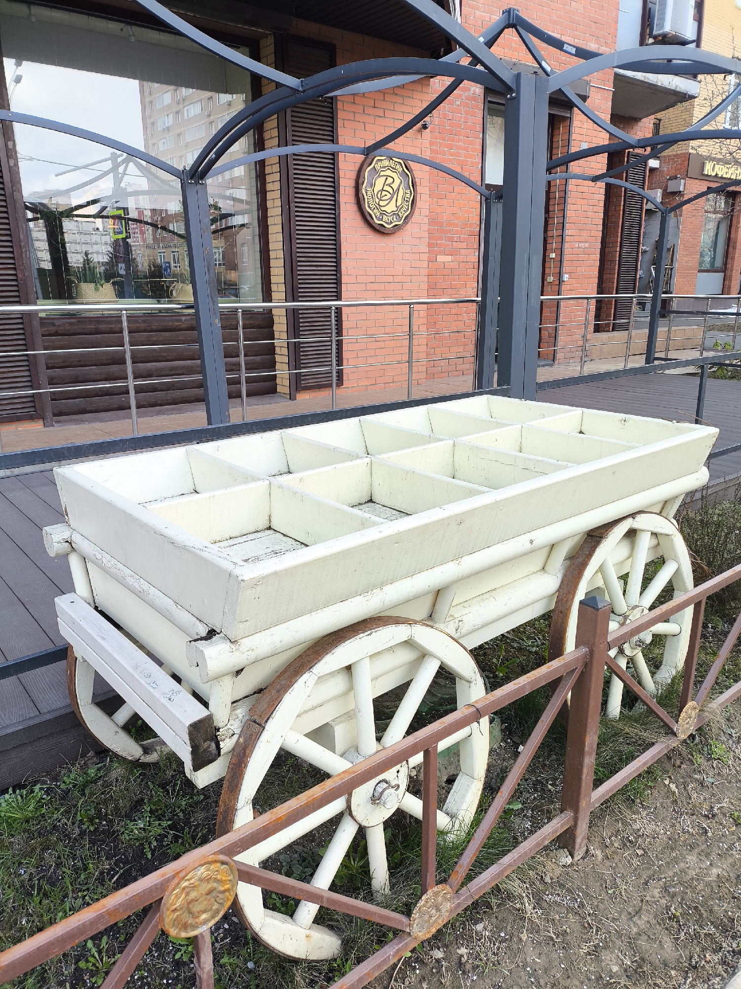 Here is a description of the image:
A decorative, white wooden cart with four wheels sits on grass next to a low, rusty metal fence. The cart is divided into sections and appears to be designed for holding plants or other decorative items. The background includes a brick building with a metal awning. The building features dark wood accents and windows. A sign with a circular emblem is visible on the brick facade. The scene is an outdoor urban environment, possibly in Moscow, Russia, considering the Cyrillic on the building's sign and architecture. There are no people visible in the immediate foreground of the scene.