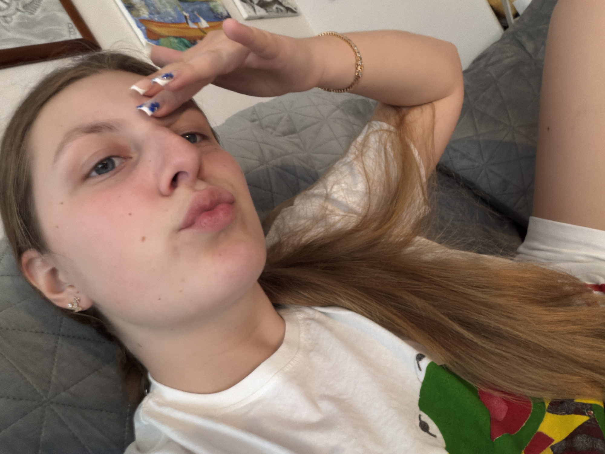 A young person with light brown hair is depicted lying on a grey quilted surface, likely a sofa or bed. They are looking towards the camera with a playful expression, featuring slightly puckered lips. One hand is raised and resting on their forehead, showcasing nails painted with a white base and blue patterns. A golden bracelet is visible on their wrist. They are wearing a white short-sleeved t-shirt with a colorful, possibly cartoon-like, design visible on the lower right, and white shorts or underwear are visible on the lower body. A small, decorative earring is worn in the left ear. In the background, framed artwork, including one with a boat scene, hangs on a wall. The lighting suggests an indoor setting during the daytime. No text is visible in the image. The location context is Burgas, Bulgaria.