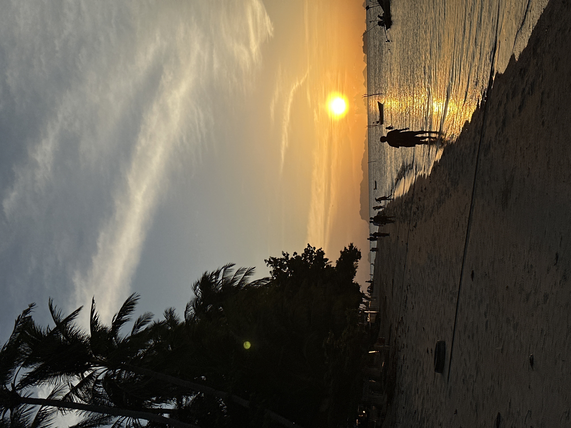 The image captures a serene sunset on the beach of Ko Samui District, Thailand. The sky is painted with hues of orange and yellow, transitioning into a soft blue, with wispy clouds streaking across. Silhouetted palm trees stand tall along the left side of the frame, their fronds rustling in the gentle breeze. The sandy beach stretches along the shore, with a few scattered objects like a discarded tire visible.
Several individuals are present, walking along the shoreline or wading into the calm, reflective water. The setting sun casts a golden glow on the water, creating a shimmering path from the horizon to the shore. Small boats are dotted across the sea, their dark shapes contrasting with the vibrant sky. The overall atmosphere is peaceful and idyllic, characteristic of a tropical beach at dusk.
