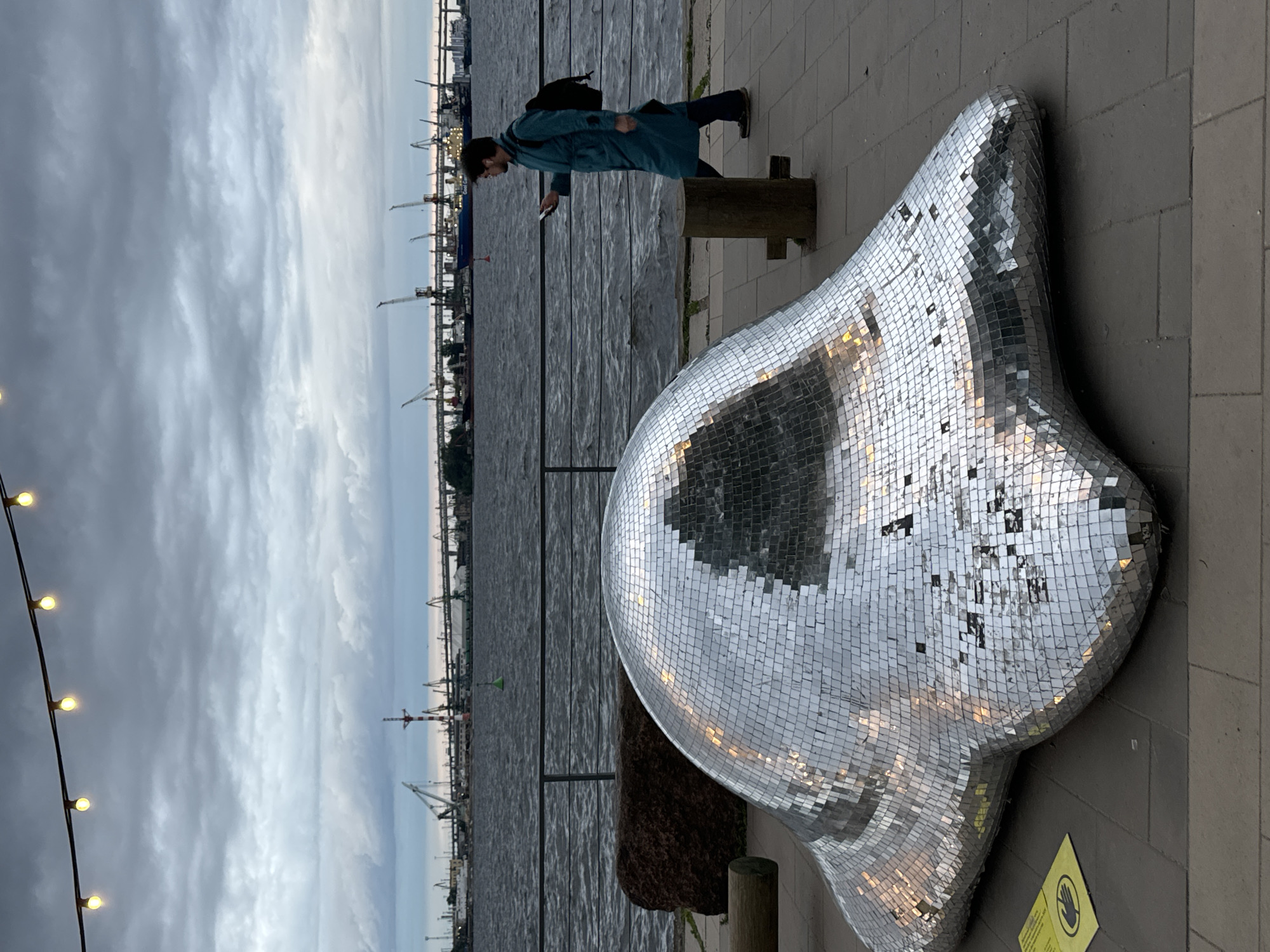 The image depicts a waterfront promenade in Saint Petersburg, Russia, on a cloudy day. In the foreground, a large, amorphous sculpture covered in mirrored tiles is situated on a paved area. To the right of the sculpture, a person wearing a teal coat and a backpack walks away from the camera, looking at their phone. They are walking on the paved promenade, which is separated from the dark, choppy water by a metal railing. In the background, across the water, is a port with numerous cranes and industrial structures. A bridge spans the water in the distance. Overhead, a string of illuminated bulbs hangs across the frame.