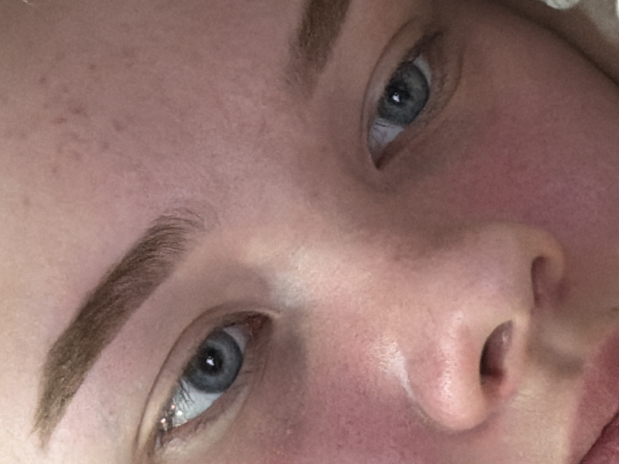 The image presents a tightly cropped, slightly downward-angled close-up of a human face, primarily focusing on the upper facial features. The subject is a single individual with fair skin exhibiting a pinkish-red flush, particularly on the cheeks and around the nasal area. The prominent features include two blue-grey eyes, with the viewer's left eye appearing more central and open, revealing a light-colored iris and a dark pupil reflecting ambient light. The right eye is visible towards the lower right portion of the frame. Both eyebrows are visible, appearing full, well-defined, and light brown/taupe in color. The bridge and partial tip of the nose are present in the lower center, and a small portion of the upper lip is visible at the extreme bottom left. The skin displays visible pores and minor textural irregularities. No specific actions or interactions are depicted. The background is entirely out of focus or beyond the frame, rendering the immediate environment indeterminate. The geographic location of the scene, including whether it is in Chaikovsky, Russia, cannot be determined from the visual content of the image.