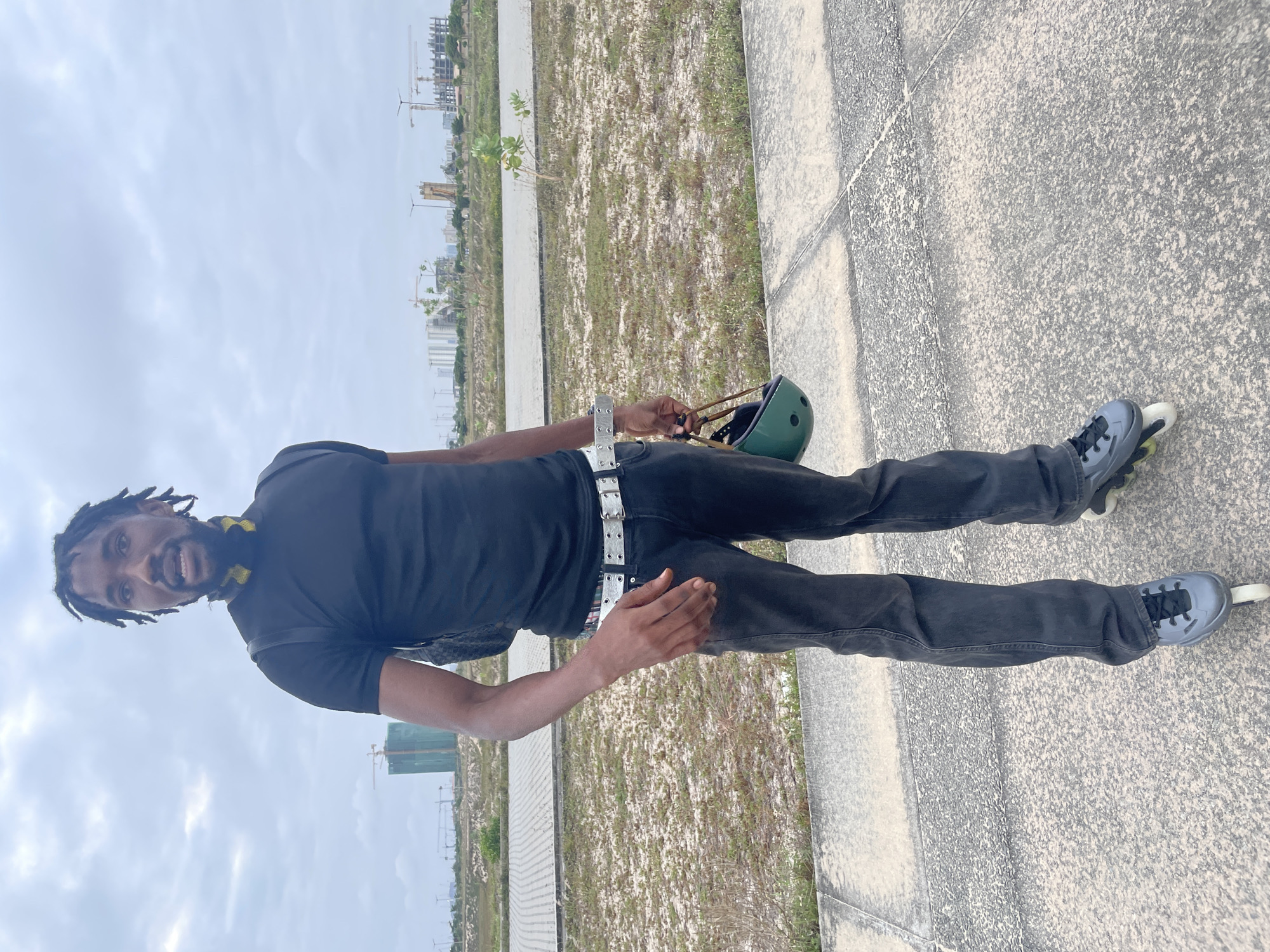 A full-body, eye-level shot captures a Black man standing on a paved walkway in Beecroft Point, Nigeria. The man, who appears to be in his late twenties or early thirties, has dark skin, dreadlocks, and a short beard. He is wearing a black t-shirt, dark gray jeans, and gray rollerblades. A green helmet is held in his right hand. In the background, construction cranes and partially built structures dominate the skyline, suggesting ongoing development. The sky is overcast and gray, and the ground is covered with a mixture of pavement and dry vegetation.
