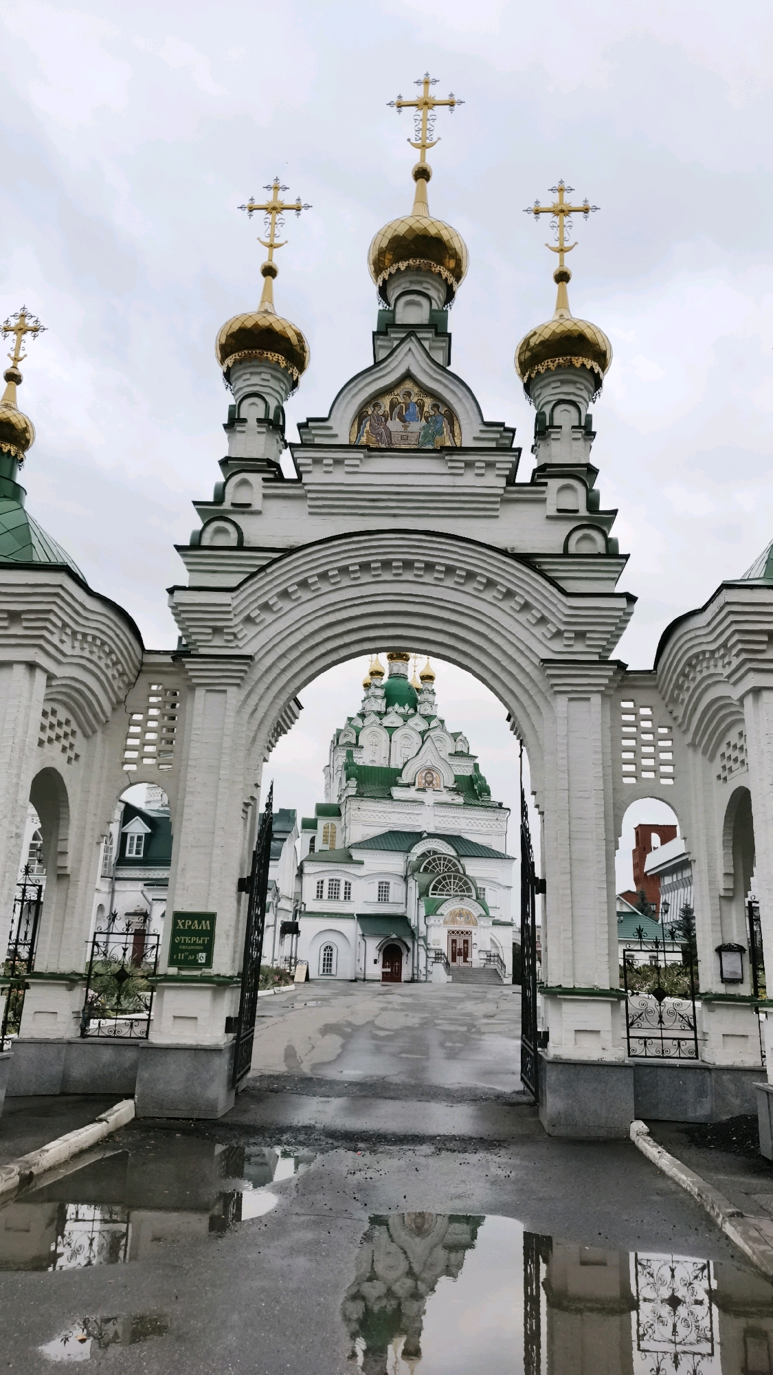 The image shows an ornate white arched gateway with a church complex visible beyond it. The gateway features decorative white brickwork and a large arch. Through the arch, a white church with green roofs and golden domes is prominently displayed. The church has multiple levels and decorative architectural elements. In the foreground, the ground is wet and reflective, showing puddles that mirror the gateway and the church. A sign with Russian text is visible on the left side of the gateway. The sky is overcast. This scene is located at the Patriarchal Square in Yoshkar-Ola, Russia, which is known for its religious architecture and themed areas.
