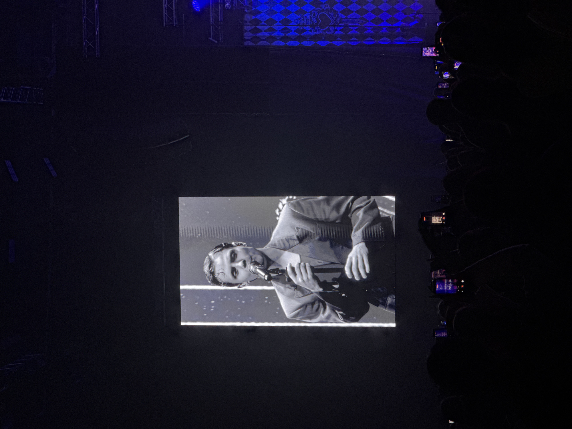 The image depicts a live music event, possibly a concert, in an indoor venue. A large screen displays a black-and-white image of a male singer holding a microphone. To the right of the screen, silhouettes of audience members are visible, many holding up phones to record the performance. Overhead, lighting rigs and a backdrop with a diamond pattern are partially visible. It is unclear if the event took place in the city of Perm, Russia, as there are no details in the picture to confirm this.