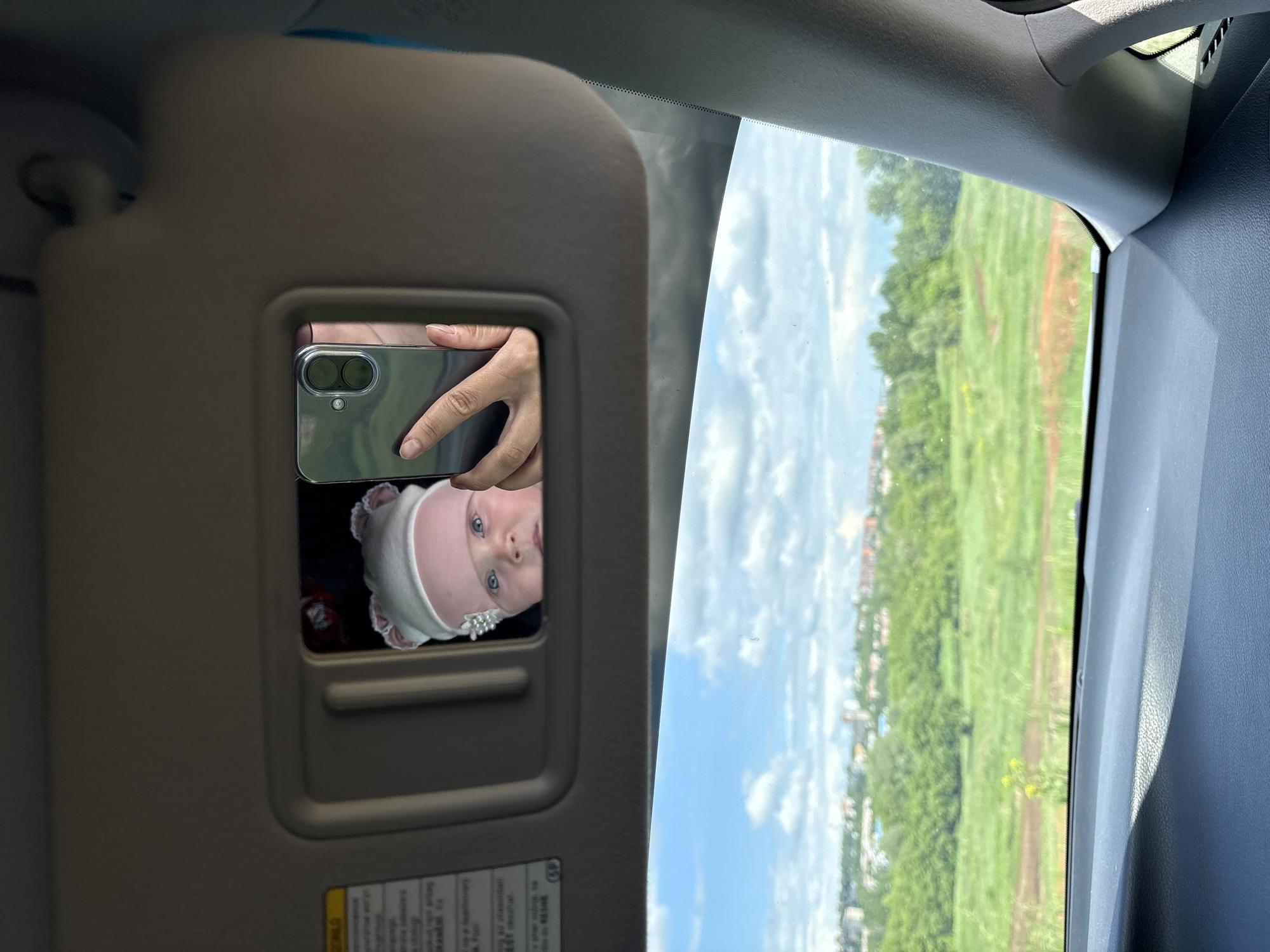 A photo taken inside a car shows a baby in a car mirror and a view of the city of Votkinsk, Russia, through the window. The baby is wearing a pink hat and is looking up at the camera. A hand is holding a smartphone to take the photo, with the reflection of the baby in the car mirror. Outside the car window is a green field with trees in the distance. The sky is blue with white clouds.