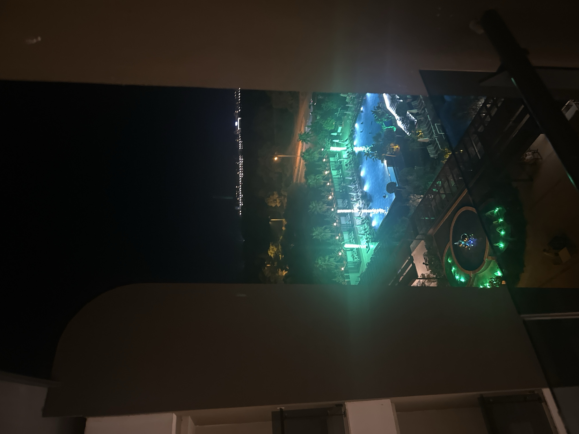 An elevated, night-time, outdoor view of a resort or hotel complex in Manavgat, Turkey, taken from a balcony or window. The foreground is dominated by dark, shadowed structures, possibly balconies or parts of the building, framing the scene.
The main focus of the image is a brightly lit swimming pool area, illuminated by vibrant green and blue underwater lights. The pool is large and rectangular, with several people visible swimming. Surrounding the pool are numerous lounge chairs and umbrellas, some of which are also illuminated. Palm trees and other lush vegetation are scattered throughout the area, many of which are also lit with green lights, creating an atmospheric glow.
Further in the background, beyond the pool, there appears to be a road with a lamppost and some distant lights of what could be a town or city skyline. The overall impression is one of a lively resort area at night, with artificial lighting creating a dramatic and colorful ambiance. A fountain with colored lights is also visible in the lower part of the image.