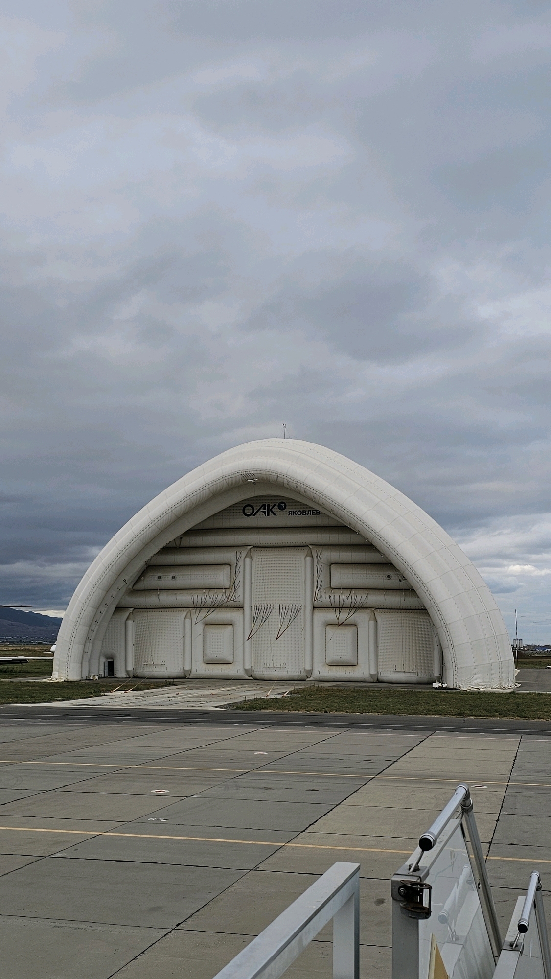 An inflatable hangar, likely an aircraft hangar, stands prominently against a cloudy sky. The hangar is white and has a curved, arched roof. Text on the facade reads "OAK" and "ЯКОВЛЕВ," suggesting it is associated with the Yakovlev design bureau, which historically produced aircraft. The hangar is situated on a tarmac, with markings indicating it's an airport or airfield. In the foreground, the edge of a gray, metal stairway or ramp is visible. The overall scene depicts an industrial or aviation-related setting in what is identified as the city of Shishkino (Domodedovo), Russia.