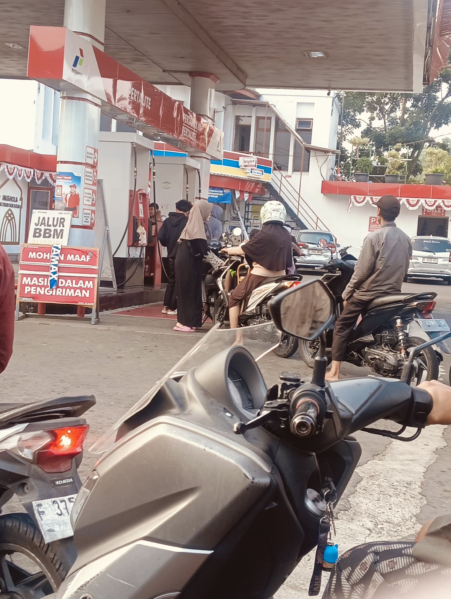 The image shows a busy scene at a Pertamina gas station in Cisarua, Indonesia. Several people on motorcycles are waiting in line to refuel. A sign in the foreground reads "JALUR BBM" (Fuel Lane) and "MOHON MAAF MASIH DALAM PENGIRIMAN" (Apologies, still under delivery), indicating a potential shortage of fuel. The gas station features red and white branding typical of Pertamina. In the background, there are several cars and buildings, including what appears to be a convenience store. The overall atmosphere is that of a normal, bustling day at a public facility.