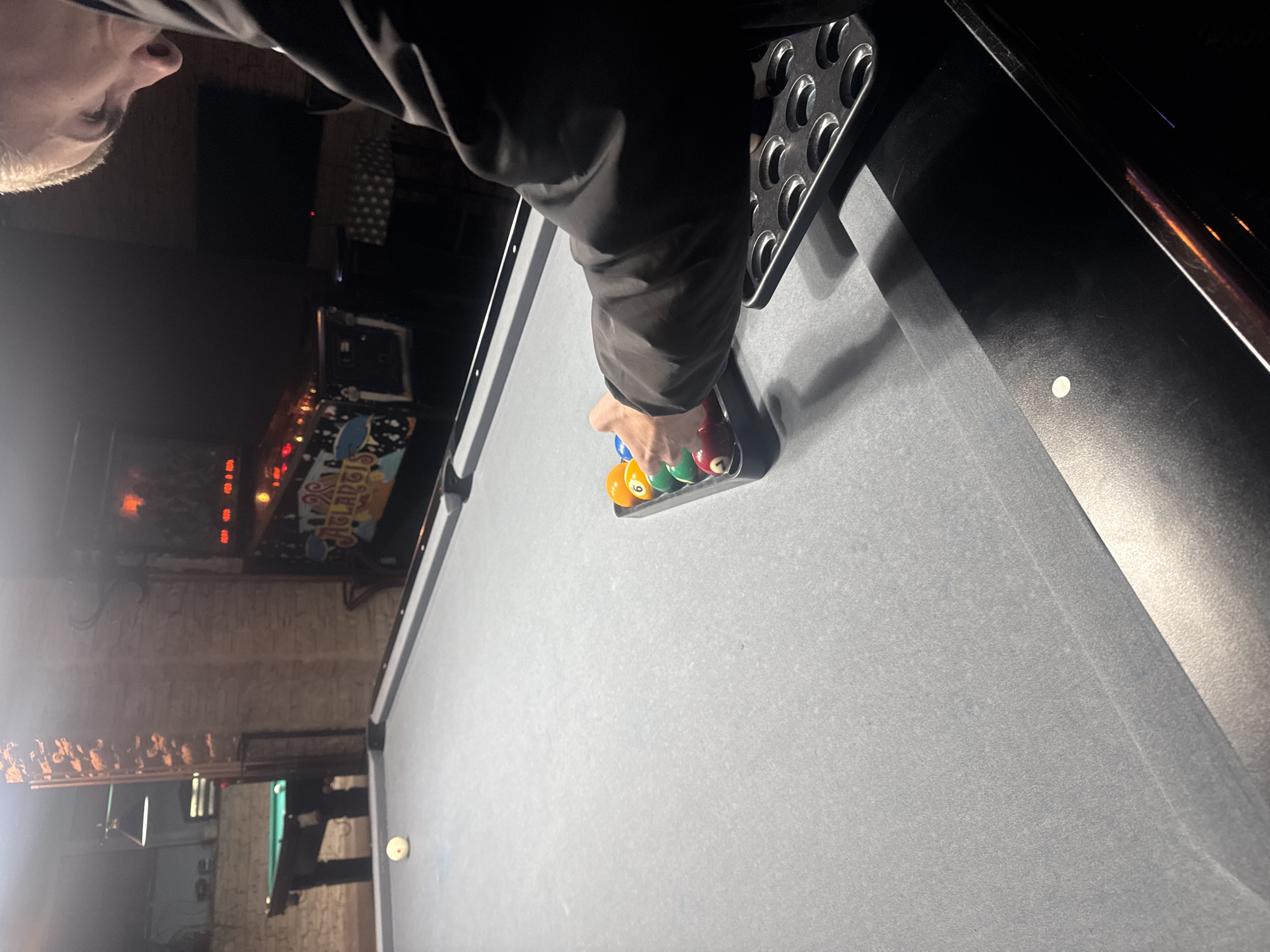 A person with short blonde hair wearing a black jacket is setting up pool balls in a triangular rack on a gray pool table. The background shows an interior space with brick walls and various light fixtures and signs. The scene is dimly lit. The location is in Plovdiv, Bulgaria.