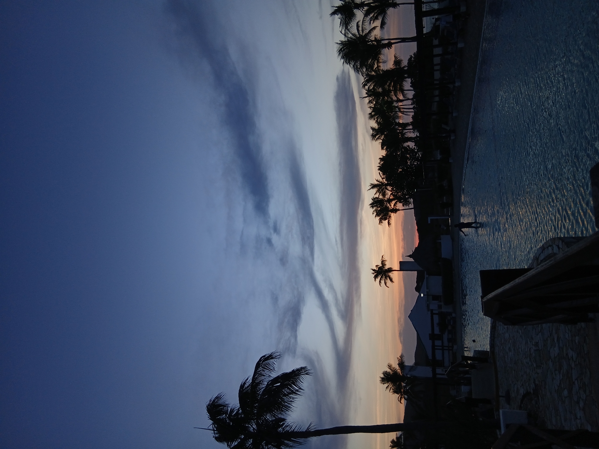 The image shows a dusk scene in Altagracia, Venezuela, featuring a swimming pool lined with palm trees. The sky displays a gradient of colors transitioning from blue to orange, with wispy clouds. Silhouettes of buildings and palm trees are visible against the fading light. A person is seen in the pool. A stone wall and wooden structure are in the foreground.