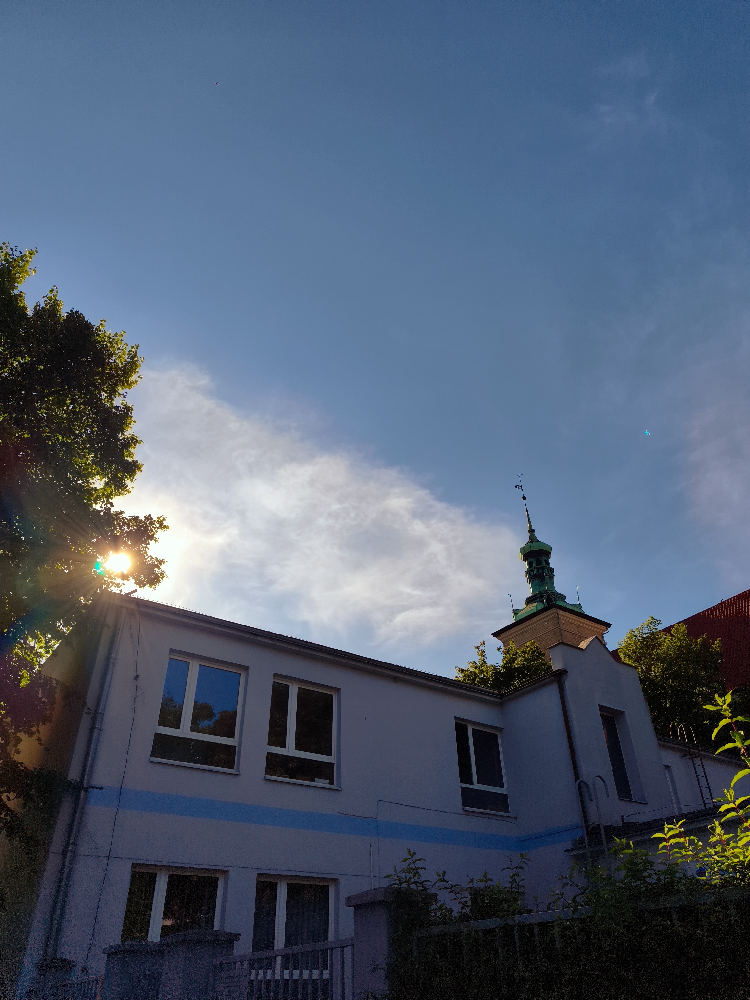 The image depicts the exterior of a building with a green-domed tower in the distance, likely situated in Gdańsk, Poland. The building is light blue with a horizontal blue stripe. It has several windows, some of which reflect the sky and surrounding trees. The sun is shining brightly, casting rays through the leaves of a large tree on the left side of the frame. The sky is clear blue with a few wispy clouds. In the far distance, a small, dark object, possibly a bird or a drone, is visible against the sky. The scene suggests a sunny day with good visibility.