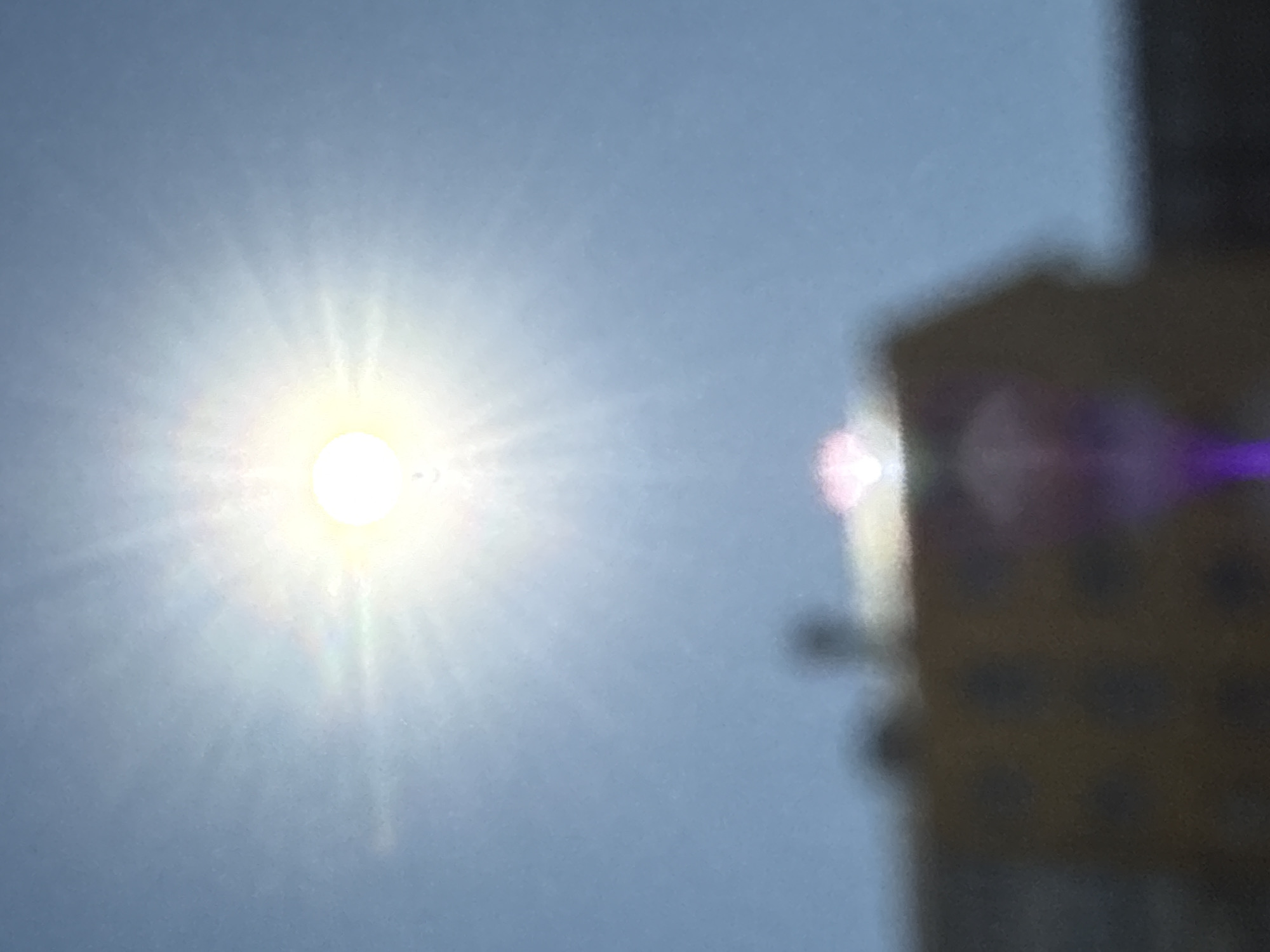 The image shows a bright sun in a light blue sky. To the right, there is a blurred yellow and dark structure, possibly a building, with a pink and purple lens flare effect on it. I cannot determine the location in Perm, Russia, or any specific details about people, subjects, objects, actions, or interactions, due to the limited context and blurred details.
