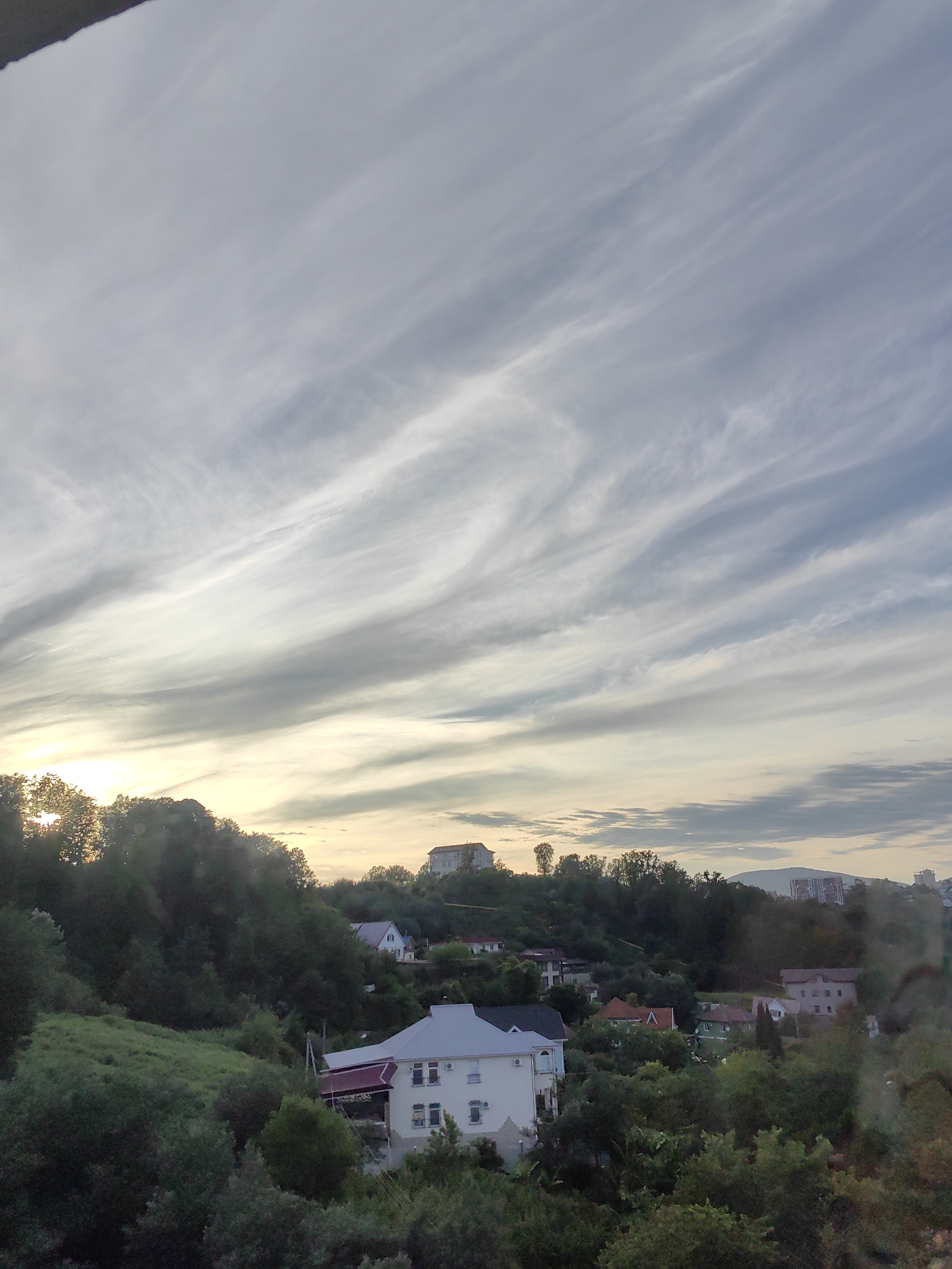 This image captures a scenic view of a hilly landscape dotted with houses, likely in Sochi, Russia. The scene is illuminated by the soft light of early morning or late evening, with the sun's rays peeking through a dense treeline on the left, creating a warm glow. Wispy, streaky clouds stretch across the pale sky, suggesting a partly cloudy or breezy weather condition.
The foreground is dominated by lush green vegetation, with trees and rolling hills that give the impression of a rural or suburban setting. Several houses are visible, varying in architectural style and color. One prominent house in the foreground is a multi-story building with white walls, a red-tiled roof, and multiple windows. Other houses, some with red and green roofs, are scattered across the hillsides, blending into the natural environment. In the distance, on a higher elevation, more buildings are visible, including apartment blocks, indicating a more urbanized area further away.
The overall mood of the image is tranquil and serene, conveying a sense of peaceful coexistence between human dwellings and nature. The natural light and the gentle undulation of the terrain contribute to a calming atmosphere. No people are visible in the frame, and there is no discernible text. The time of day is suggested by the low angle of the sun and the soft, diffused light.