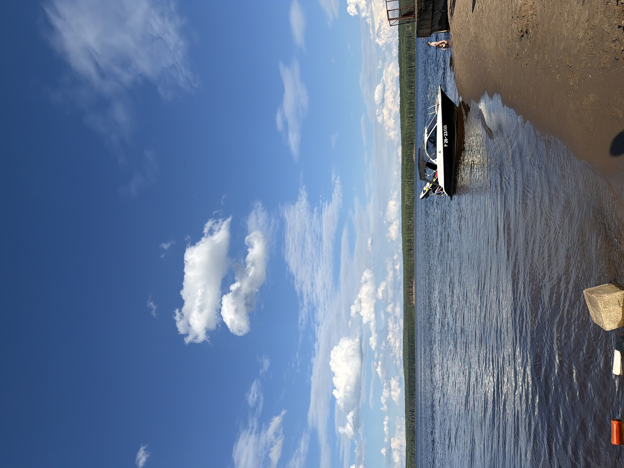 This is a portrait-oriented photograph taken on a sunny day, likely in the late morning or early afternoon, given the bright illumination. The scene depicts a body of water, possibly a river or lake, with a sandy shore on the right. The water is calm with gentle ripples reflecting the sunlight, suggesting fair weather.
Dominating the left side of the frame is a vast expanse of blue sky dotted with fluffy white clouds. The clouds range from wispy cirrus formations to larger cumulus clouds, creating a picturesque sky.
Along the shoreline, a modern-looking motorboat is partially beached. It is black and white with a black canopy. The boat has a license plate or registration number visible on its side, reading "P2B-23111". Behind the boat, a small person is visible in the water near a pier or structure extending into the water. Further back, a dense line of green trees stretches along the horizon, indicating a natural, possibly forested, landscape.
On the sandy shore, a large, light-colored rock is prominently placed in the lower right corner, along with a couple of smaller, lighter-colored objects, one of which appears to be a white buoy or marker, and another is orange. The overall mood of the image is serene and leisurely, capturing a pleasant day by the water. The location context suggests this scene is in Chaikovsky, Russia.