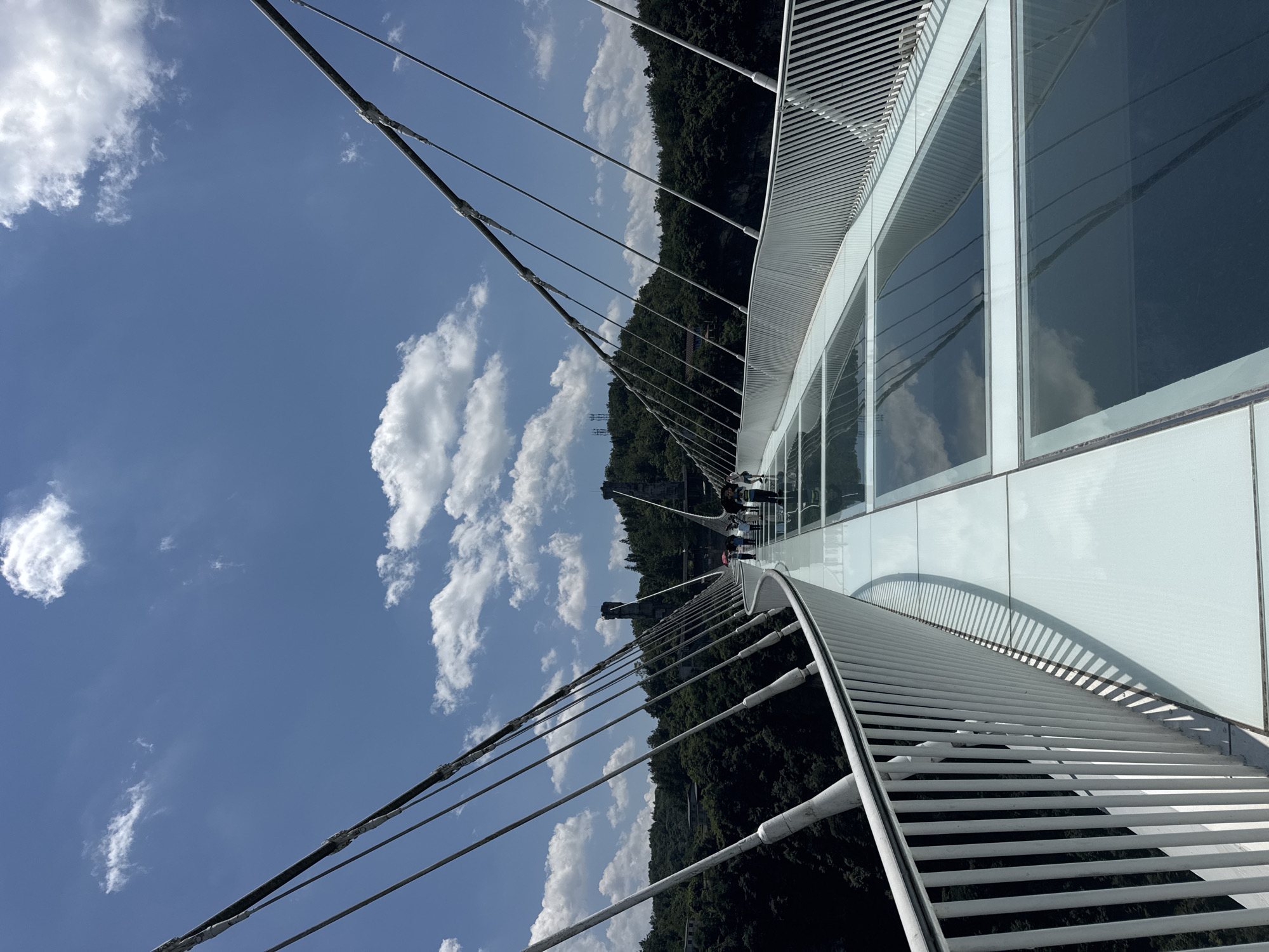 The image shows a perspective view of a glass-bottomed suspension bridge. The bridge has white railings on both sides and is supported by numerous thick cables that stretch upwards and outwards, disappearing into the distance. The bridge floor is made of large, rectangular glass panels, reflecting the blue sky with scattered white clouds. Several people, some with umbrellas, are walking along the bridge in the distance, appearing as small figures against the vast landscape. The bridge spans across a deep gorge, with lush green, tree-covered mountains rising steeply on either side. In the background, two tall, stone-like structures with platforms at their tops are visible, possibly observation towers or part of the bridge's support system. The scene is set in a natural environment under a bright, sunny sky. This bridge is likely the Zhangjiajie Grand Canyon Glass Bridge in Zhangjiajie, China, known for its similar design and surrounding mountainous terrain.
