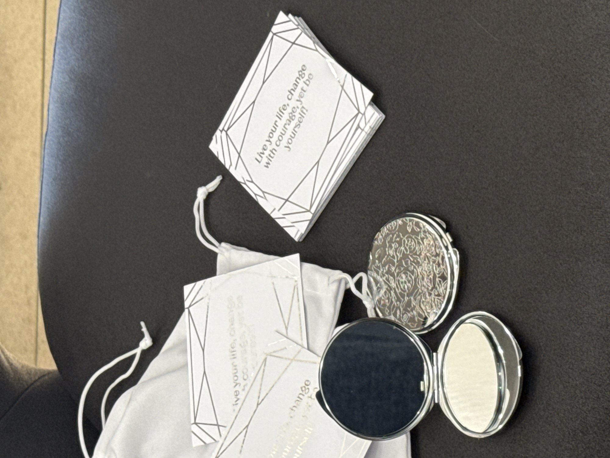 The image shows two compact mirrors and several cards with text and geometric designs, laid out on a dark, textured surface. A white drawstring bag is also visible. One compact mirror is open, revealing a reflective surface. The other compact mirror is closed and features an embossed floral pattern. The cards appear to be quote cards, with the visible text reading "Live your life, change with courage, yet be yourself!". The scene is lit from above, casting subtle shadows. There is no indication of the city of Sunzha, Russia.