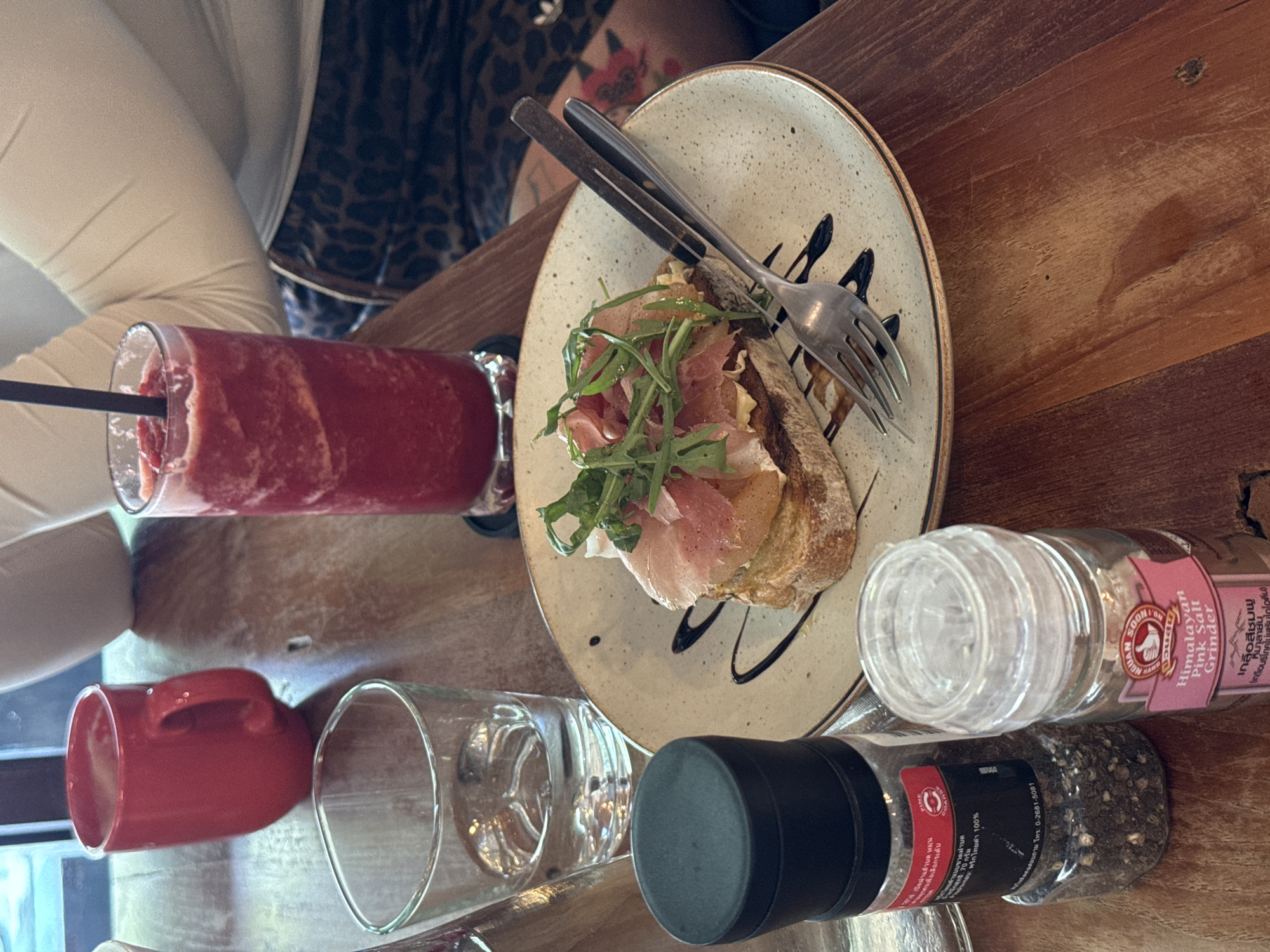 Here is a description of the image:
The image shows a table setting in what appears to be a restaurant or cafe, given the variety of food and beverage items. There's a plate with a slice of bread topped with cured meat, some greens, and drizzled with a balsamic glaze. A fork and knife are laid across the plate. To the left, there's a tall glass of red smoothie, next to a red cup and an empty glass. To the right of the plate are grinder jars for salt and pepper. The label on the Himalayan Pink Salt grinder is in Thai. The image is taken from an overhead angle, showcasing the wooden texture of the table.
Given the Thai label and the style of food, it is possible this scene is located in Chiang Mai City Municipality, Thailand.