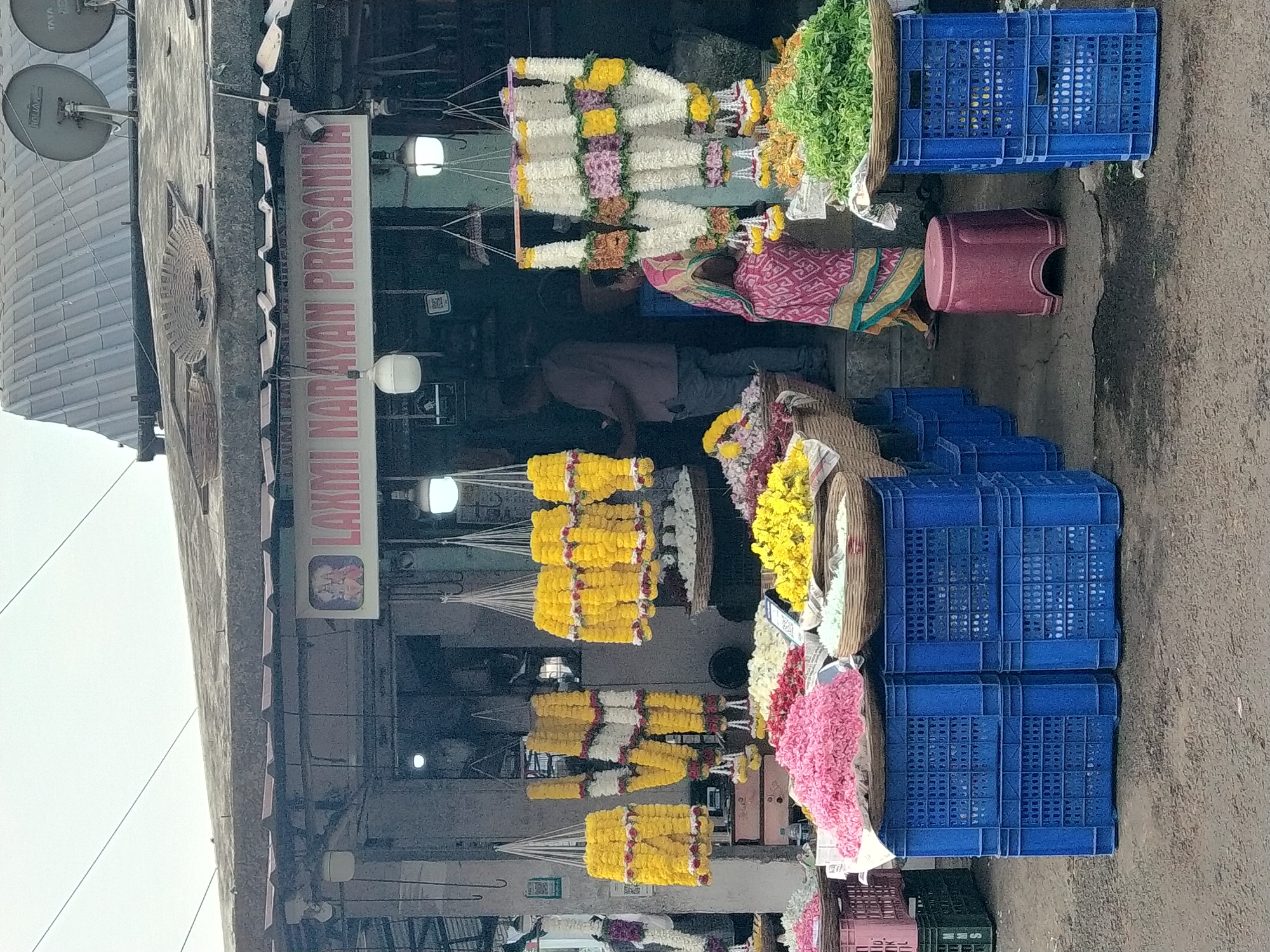 Here's a description of the image:
The image shows a flower market storefront in Panaji, India. The storefront has a sign that reads "Laxmi Narayan Prasanna" with what appears to be images of Hindu deities. Rows of strung garlands hang from the ceiling, comprised of various flower types and colors, including yellow, white, pink, and multicolored combinations. These garlands are positioned in front of the store, accessible from the street. Green foliage is visible among the flowers.
A person with a colorful shawl or garment stands inside the storefront area. There are other figures partially visible inside the store.
To the right of the storefront, there are several stacked blue plastic crates, some containing flowers or foliage. A pink stool is visible near the crates. The ground appears to be paved. Satellite dishes are mounted on the building’s roof.