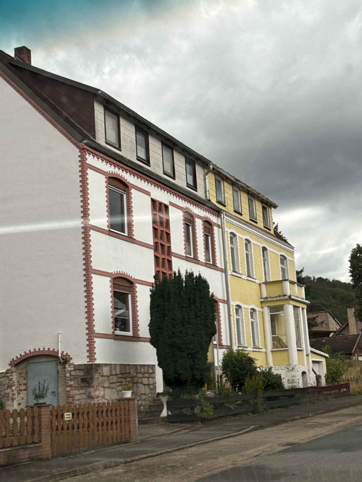The image depicts a row of buildings in the city of Bad Sachsa, Germany, under a cloudy sky. On the left, a white building with red brick accents features several windows. A wooden fence with vertical slats runs along the front of this property. To its right, a bright yellow building is visible, also with multiple windows and architectural detailing. A dense evergreen tree stands in front of the yellow building. The background shows a hillside covered in trees. The scene appears to be a residential area, with no people or specific activities immediately apparent.