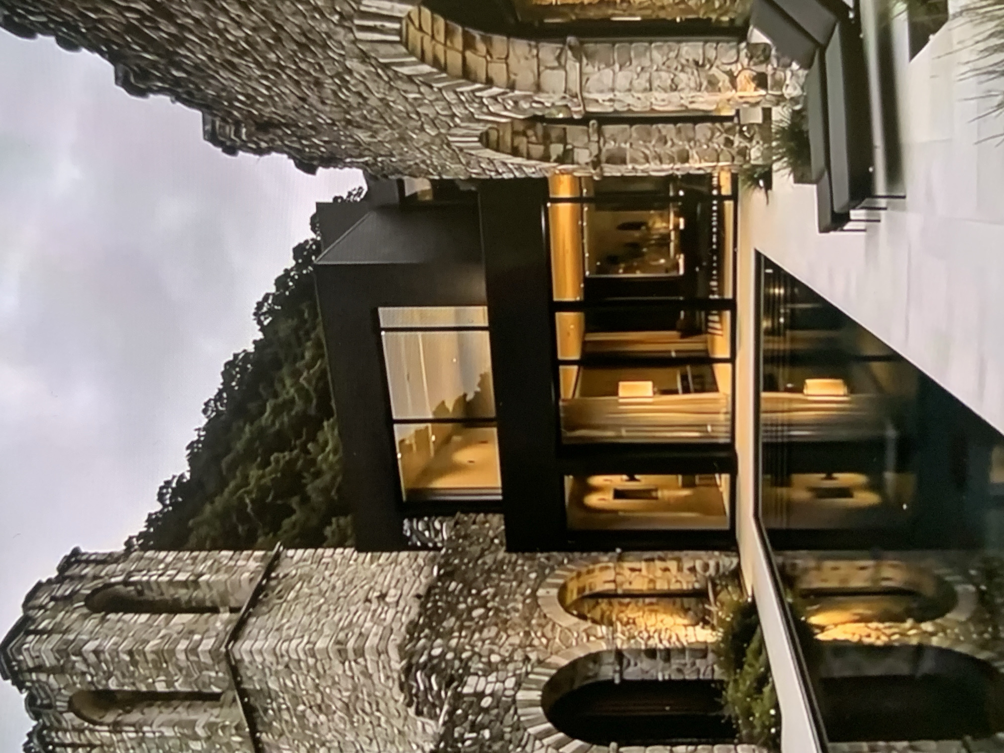 The image depicts a modern black house integrated with ancient stone structures, featuring arched openings and a tower-like section on the left, and a ruined stone wall with arches on the right. A swimming pool is situated in the foreground, reflecting the illuminated interiors of the house and the stone architecture. The house has large glass windows and doors, revealing warmly lit living spaces with furniture. Two dark lounge chairs are placed on the white tiled patio next to the pool. Behind the structures, a densely forested hillside rises under a cloudy sky. The scene appears to be a fusion of historical and contemporary design in an outdoor setting, likely a private residence. The information about Stavropol, Russia, is not visually supported by the image itself.