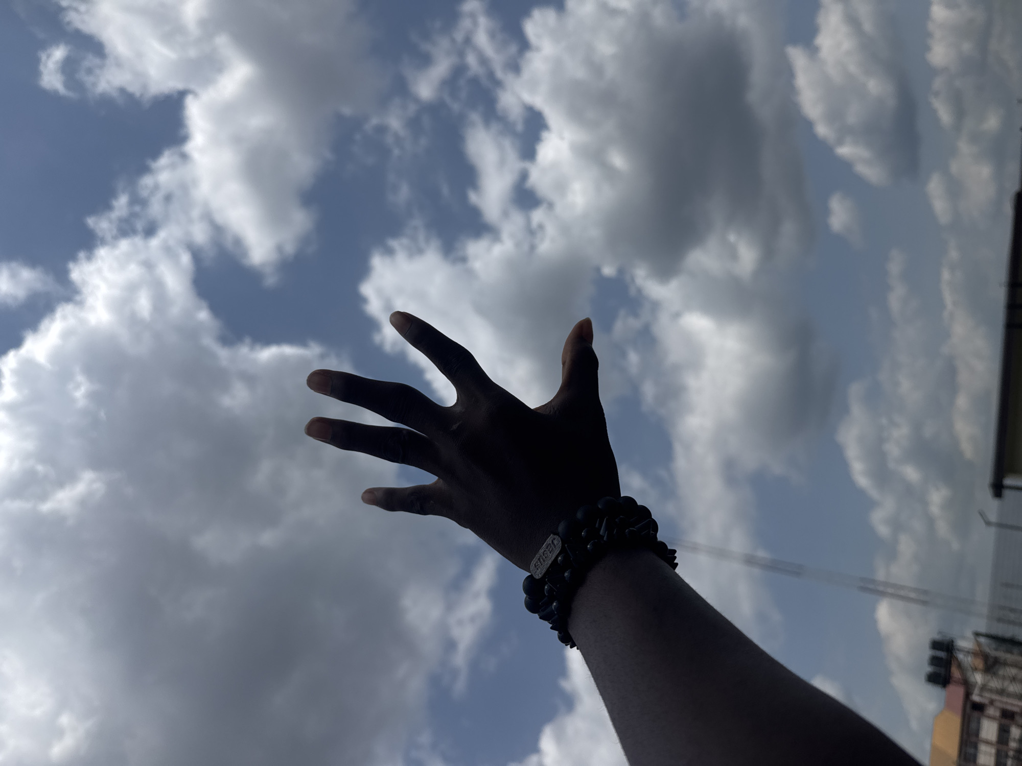 The image captures a human left arm and open hand, silhouetted against a partly cloudy sky, with fingers spread and reaching towards the upper right. Multiple black beaded bracelets are worn on the wrist, one featuring a metallic rectangular charm with the word "JESUS" visible. The background consists of a blue sky largely covered by white and grey cumulus clouds. In the lower left, indistinct distant structures are visible, consistent with an urban environment in the city of Ihiagwa, Nigeria, including a building with an orange-red roof, black water tanks on its upper portion, and a blurred, tall communication tower.