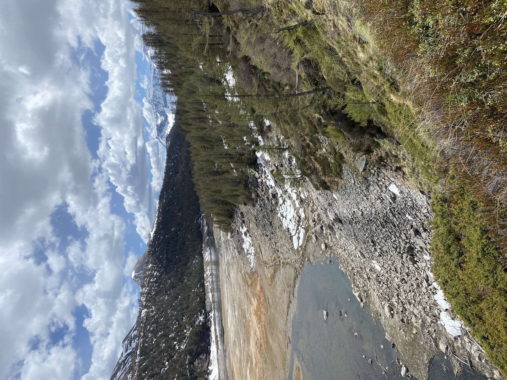 The image depicts a landscape near Baceno, Italy, featuring a partially dried-up lake or reservoir. The foreground contains a body of water with exposed lakebed areas of dry, cracked sediment. Rocks and patches of snow are visible along the water's edge and on the slopes leading down to the lake. A densely wooded hillside with green trees borders one side of the lake. In the background, a mountain range with snowy peaks is visible under a blue sky dotted with fluffy white clouds.