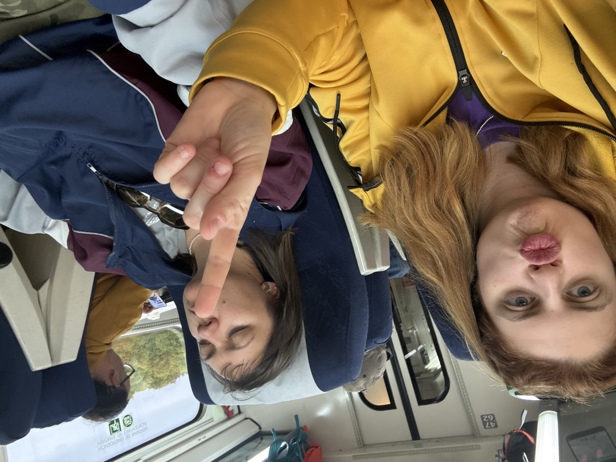 Two women are seated on a train, with one woman asleep and the other making a puckered face for a selfie. The woman on the left is sleeping with her eyes closed, and one hand raised making a peace sign near her face. She has on a necklace. A blue jacket lies across her lap. The woman on the right has shoulder-length, light brown hair and is wearing a yellow jacket. In the background, a man can be seen looking out the train window. The window shows passing trees. The train car has blue seats and white walls. The train's window has an emergency exit symbol printed on it. The location is identified by the text on the window, indicating a railcar from RENFE, the Spanish National Railway Network. It is assumed to be in Spain, since RENFE is the state-owned Spanish company. The train's car numbers appear on the bottom right of the window. It is not possible to confirm if it is near Sarria, Spain, since there are no indicators of that city.