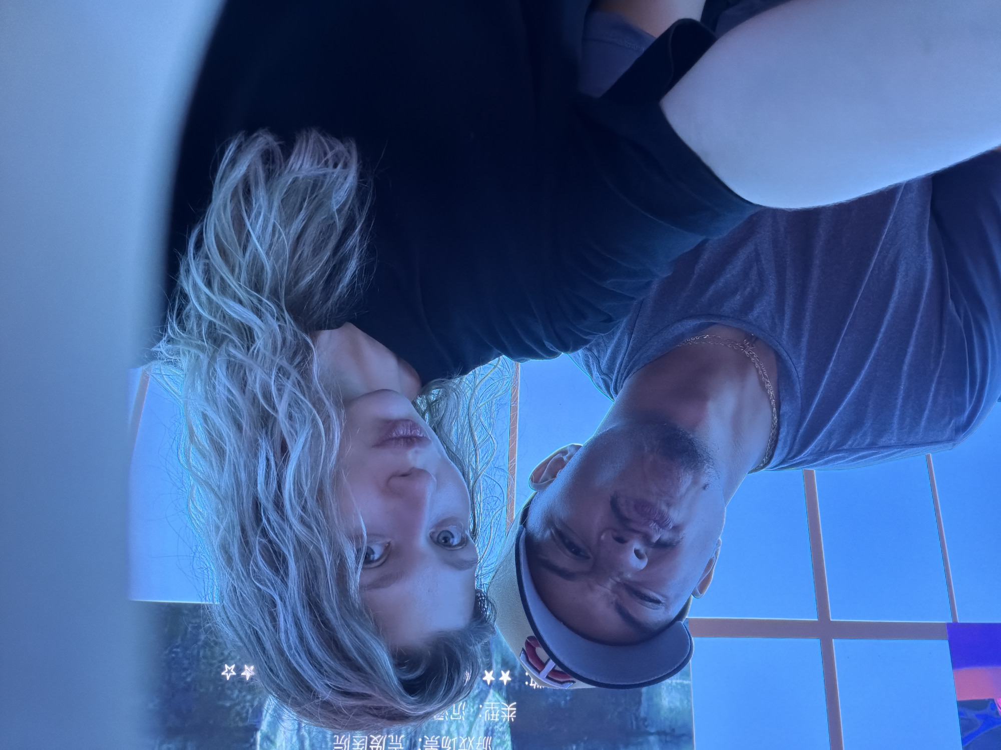 A selfie is taken by a young man and woman in a dimly lit room. The man is on the left, wearing a grey t-shirt and a baseball cap, making a funny face. The woman is on the right, with blonde, curly hair, wearing a black t-shirt and posing with a pout. They are likely in a public space with blue lighting, possibly a game arcade or entertainment venue, as a screen displaying Chinese text and stars is visible in the background. The scene suggests a casual and playful moment captured in Dongguan, China.