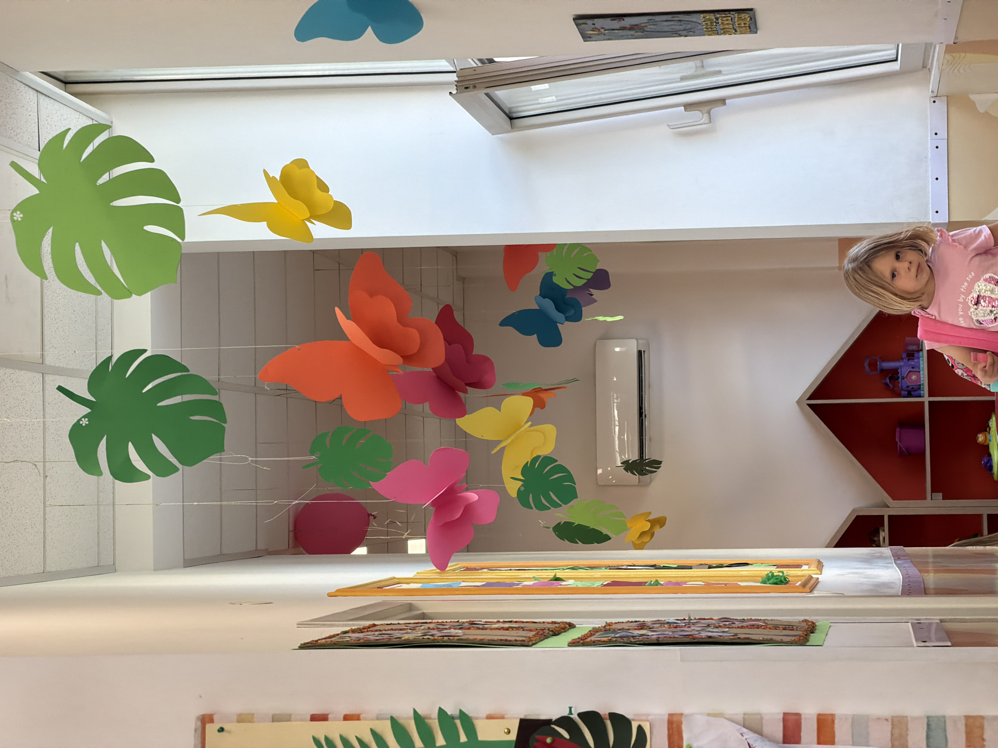 A low-angle, indoor shot displays a room decorated with paper tropical leaves and colorful paper butterflies suspended from the ceiling by clear string. A young, fair-skinned girl with blonde hair is visible on the right side of the frame, peering into the room. She is wearing a pink t-shirt.
Visible within the room are shelves filled with children’s toys, and an air conditioning unit mounted on the wall. Artwork, possibly children's crafts, are positioned in the foreground. A skylight is visible in the ceiling. Based on the observed features typical of children's play areas, it is inferred the location could be a kindergarten or daycare setting within Budva, Montenegro.