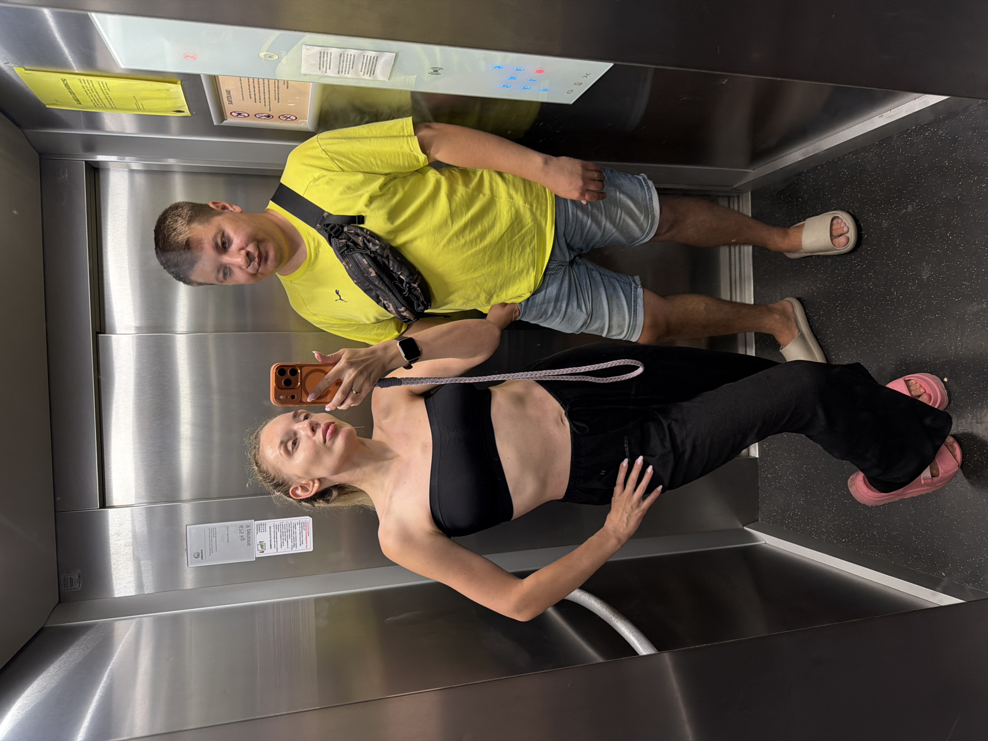 An elevator in Budva, Montenegro, contains a man and a woman posing for a mirror selfie. The woman, on the left, wears a black bandeau top and black pants, with bright pink slides. She holds an orange-cased smartphone. The man, on the right, wears a yellow t-shirt and denim shorts, with beige slides and a black fanny pack across his chest. The elevator walls are made of brushed metal, and there are various signs and buttons visible on the walls. The floor is dark and speckled.