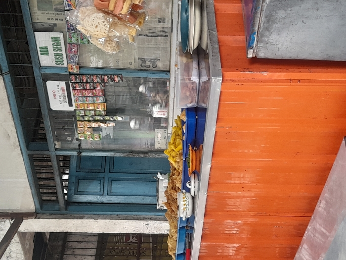 The image displays a food stall located in Pandanarum, Indonesia. A central orange-paneled counter, topped with a metallic surface, presents a variety of food items. Multiple blue plastic trays contain assorted fried foods, including fritters and what appear to be fried cassava or potato wedges. Several ceramic bowls, one with a lid, and a small dish are arranged on the counter. A clear plastic container holds more food, and stacked plates are visible on the right. In the background, a building facade features a large window section and a closed blue wooden door. The window displays small packaged items hanging, and two white signs are affixed: "INFO KEDAI INI SELASA LIBUR" (This stall is closed on Tuesdays) and "ADA SUSU SEGAR" (Fresh milk available). Reflections on the window glass obscure visibility beyond, but blurred shapes consistent with a motorcycle or scooter and exterior structures are discernible. Above the window and door, dark horizontal bars are present. A partial red sign displaying "SAMPOERNA" is visible on the far left, behind a concrete pillar. No people are directly depicted in the scene.