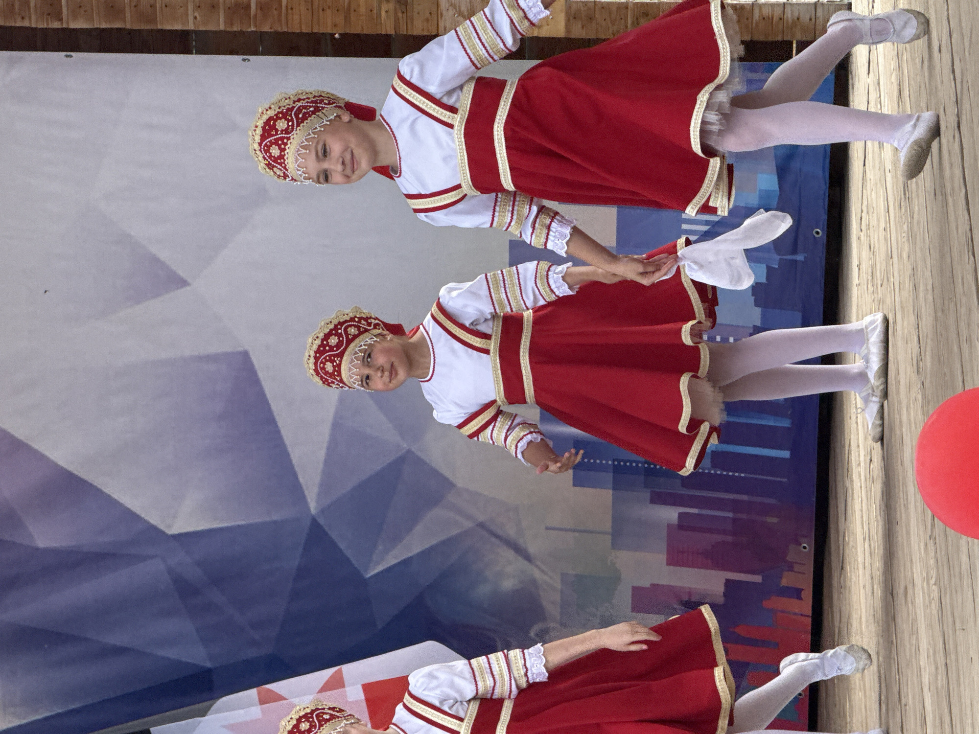 In Votkinsk, Russia, on a wooden stage with a backdrop featuring a geometric pattern and a city skyline illustration, three young girls in traditional Russian costumes perform. The girls, who appear to be of similar ages, are dressed in matching red dresses with white sleeves and elaborate gold and red headpieces. The girl on the right is smiling and looking towards the audience, while the girl in the middle is also smiling and holding a white cloth. The girl on the far left, partially visible, is also in costume. The background consists of a large banner with a blue and white abstract design on the left, and what appears to be wooden siding on the right. A large red ball is visible in the foreground.